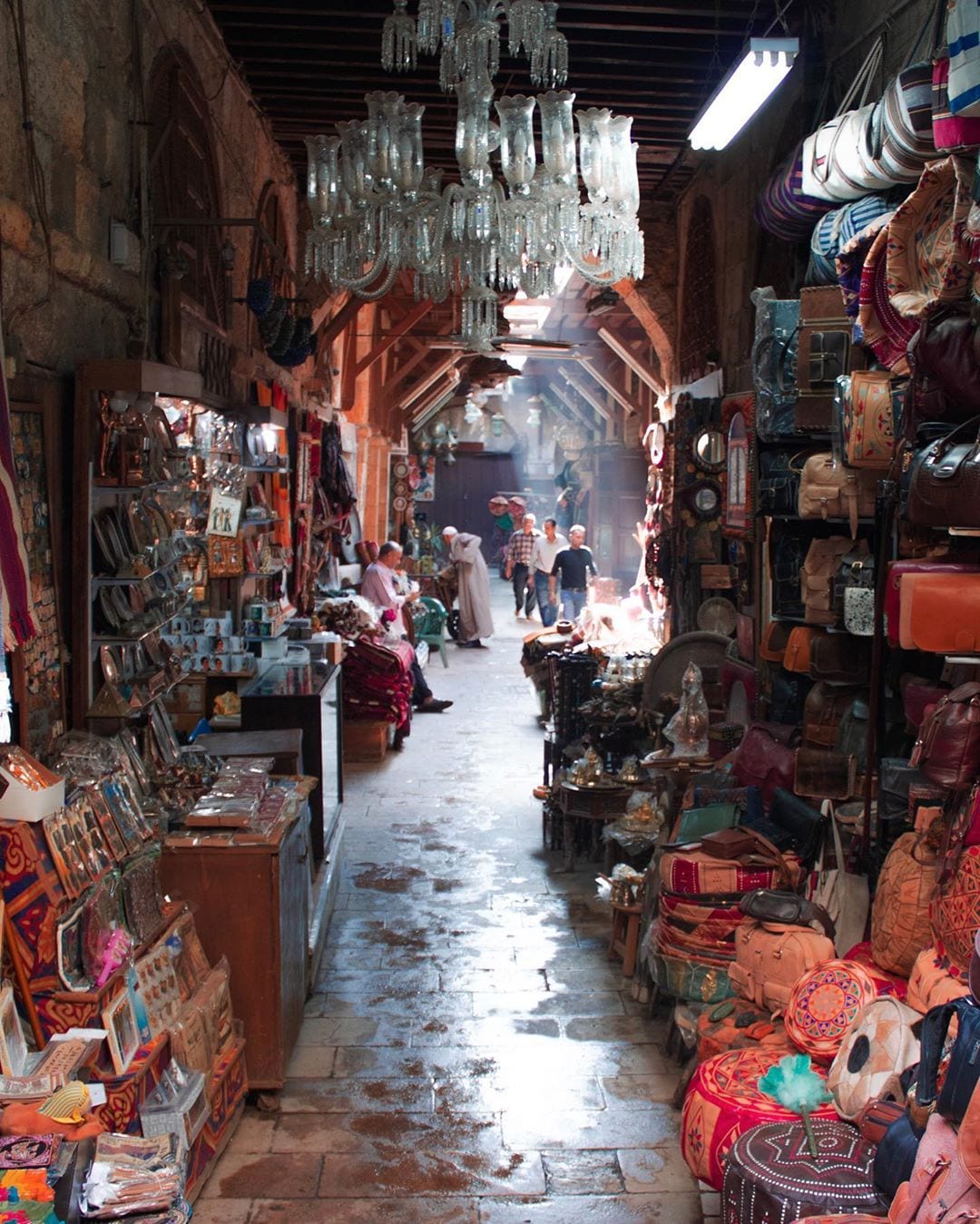 A narrow covered market alley hung with leather bags, cushions and lamps, with vendors and shoppers walking through