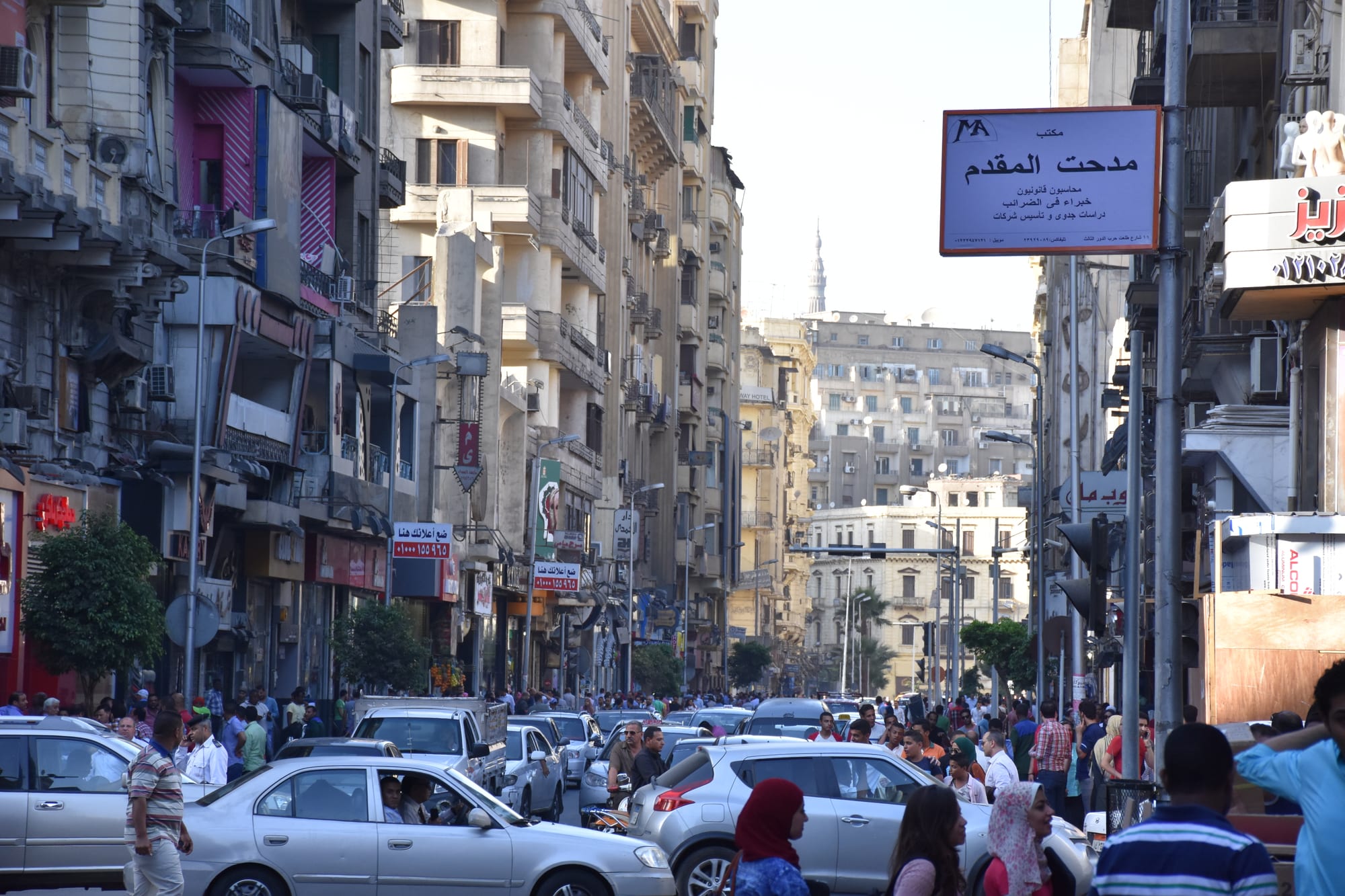 A crowded Cairo street packed with cars, motorbikes and pedestrians between tall apartment buildings, a minaret visible in the distance