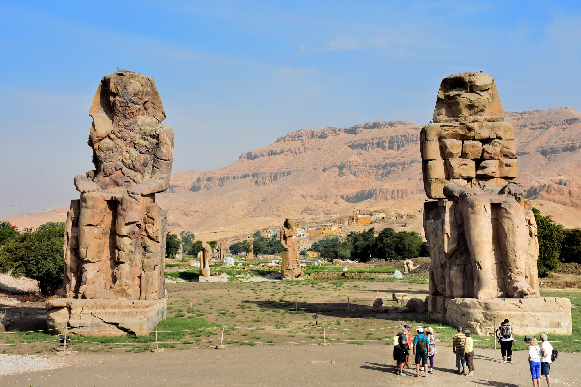 Two colossal weathered stone statues of seated figures facing the camera across open ground, with green fields and bare desert cliffs behind them