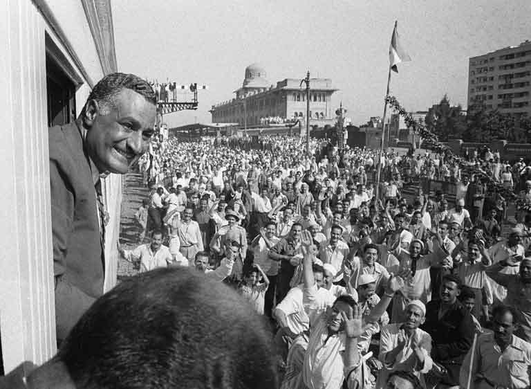 Gamal Abdel Nasser standing in an open car surrounded by a dense, cheering crowd