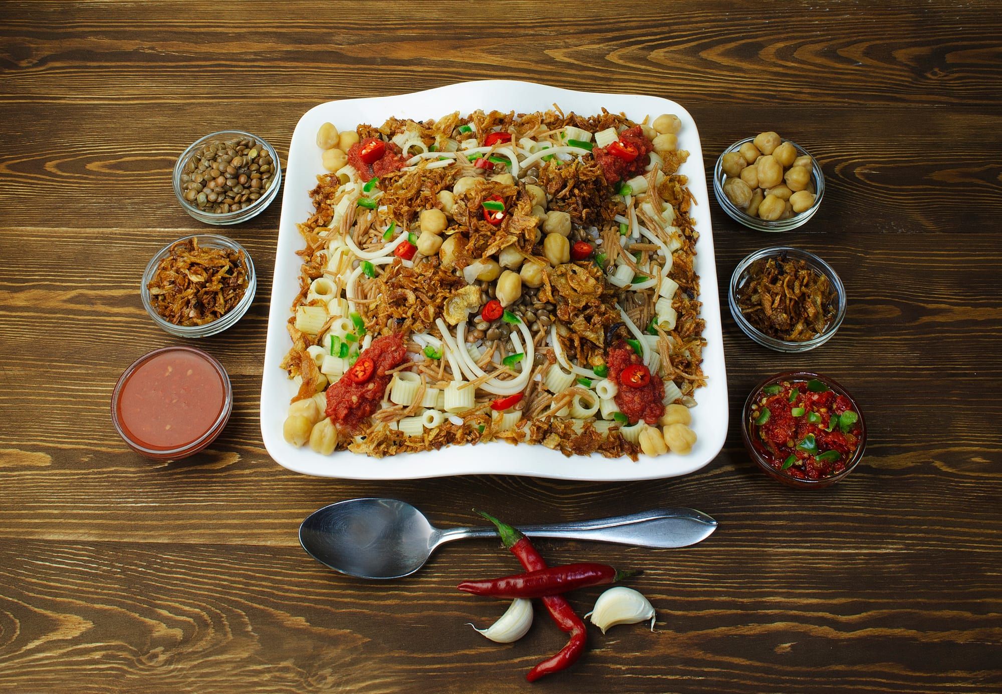 A bowl layered with rice, lentils, pasta and chickpeas, topped with fried onions and a red sauce
