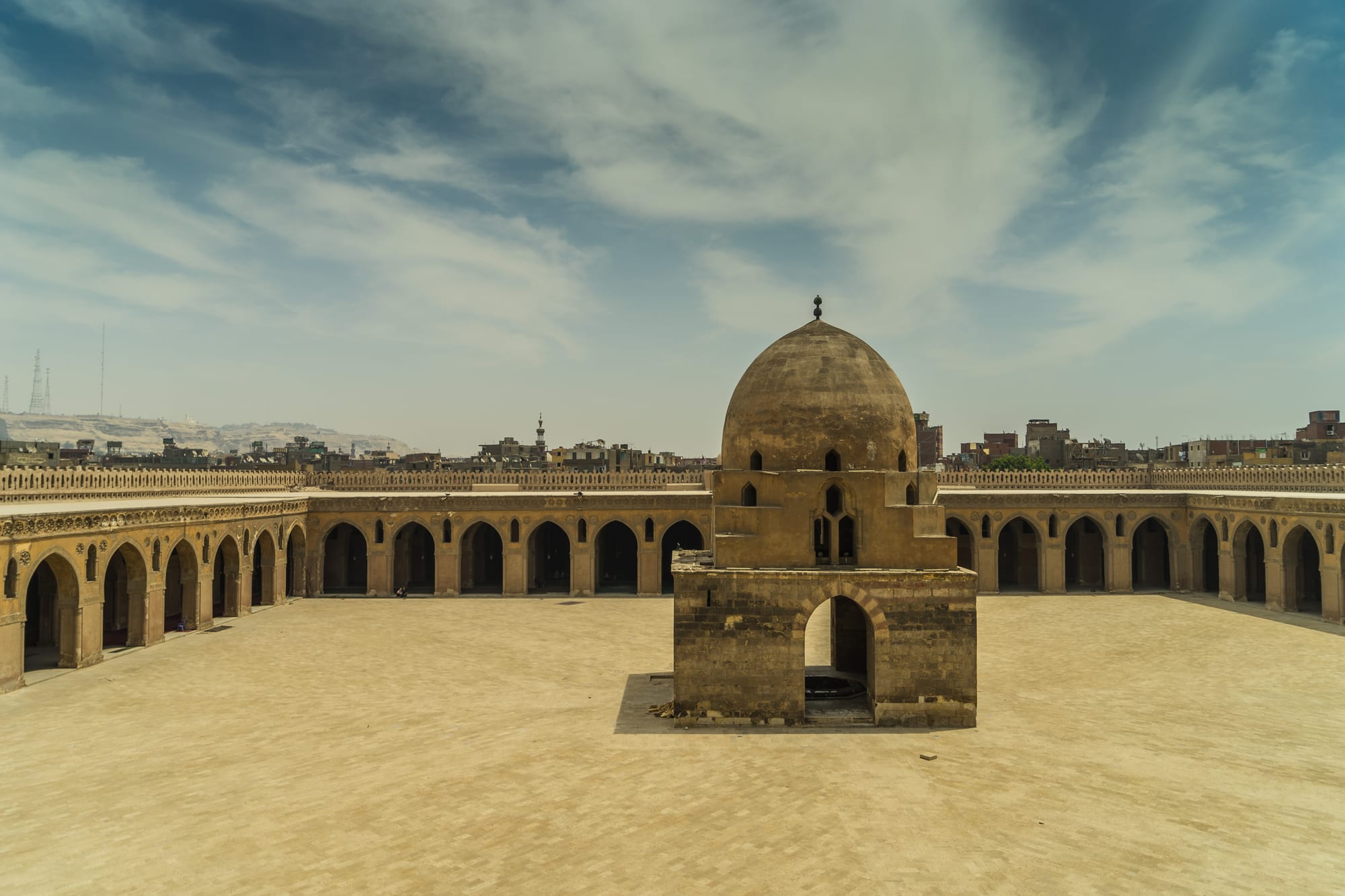 A vast, empty sandy-coloured courtyard surrounded by arcades of pointed arches, with a square domed fountain building at its centre under a pale blue sky