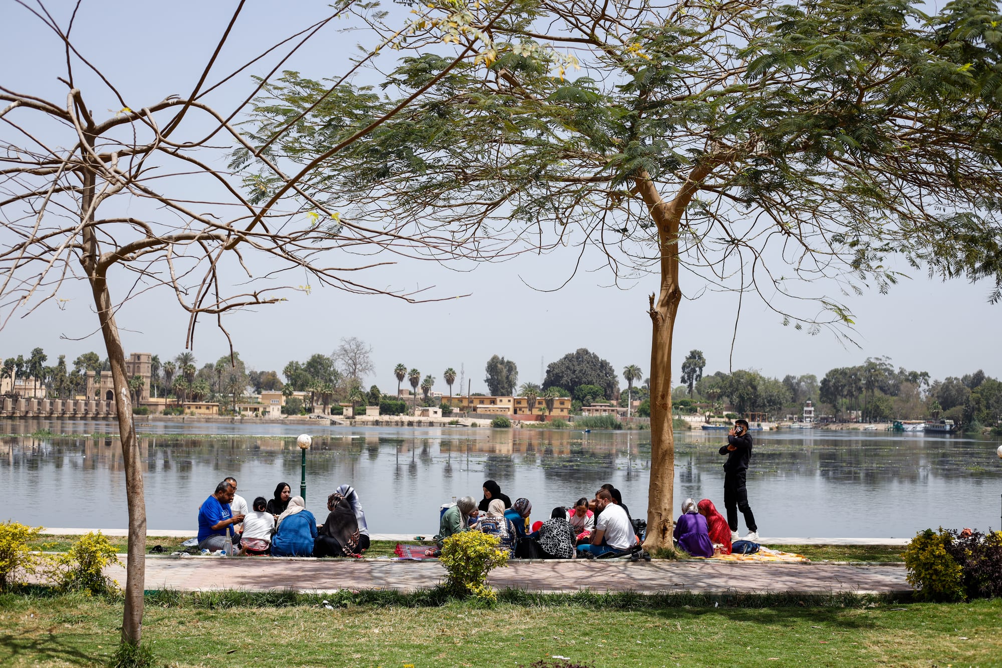 Several groups of people sit on blankets on a paved riverside path under trees, with a wide calm river and palm trees on the far bank