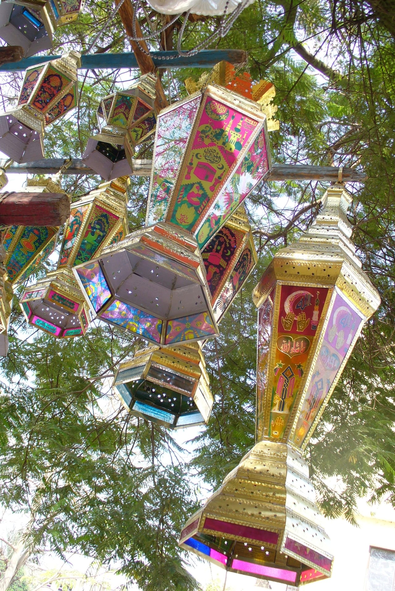 Looking up at a cluster of large, brightly coloured metal-and-glass lanterns with star-shaped cut-outs and printed patterns, hanging from wooden poles among the branches of a tree