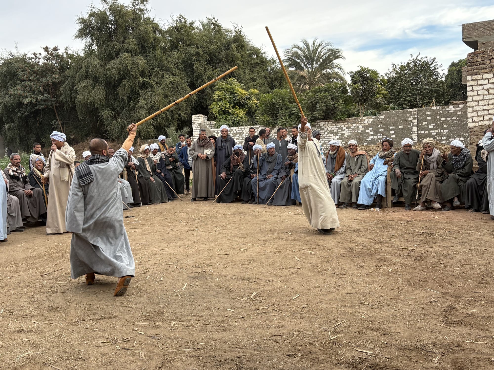 Two men in long robes face each other on a dusty open ground, each raising a long wooden stick, watched by a large semicircle of men in robes and turbans seated and standing along a wall