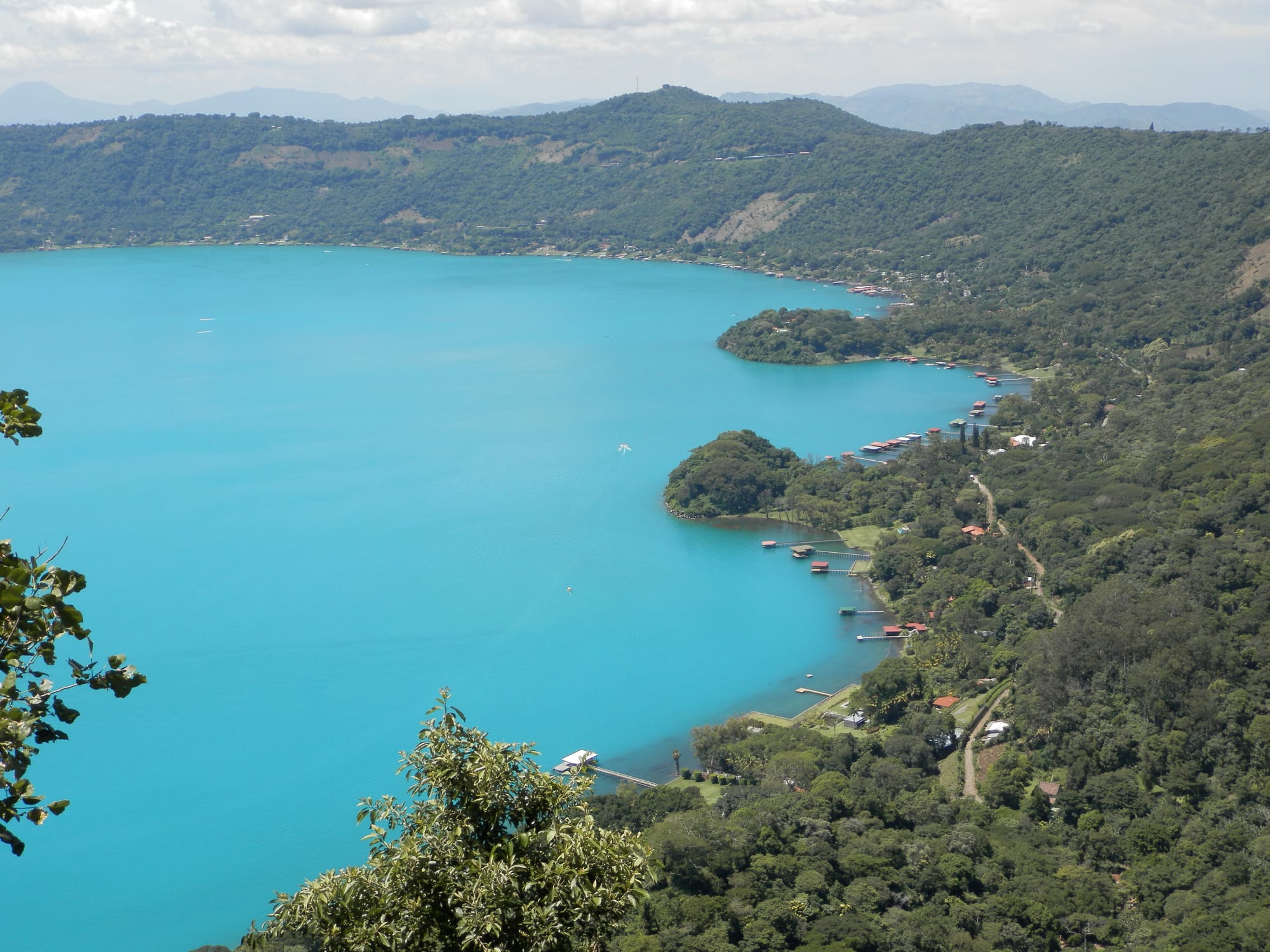 A large round crater lake of deep blue-green water ringed by forested hills and small houses