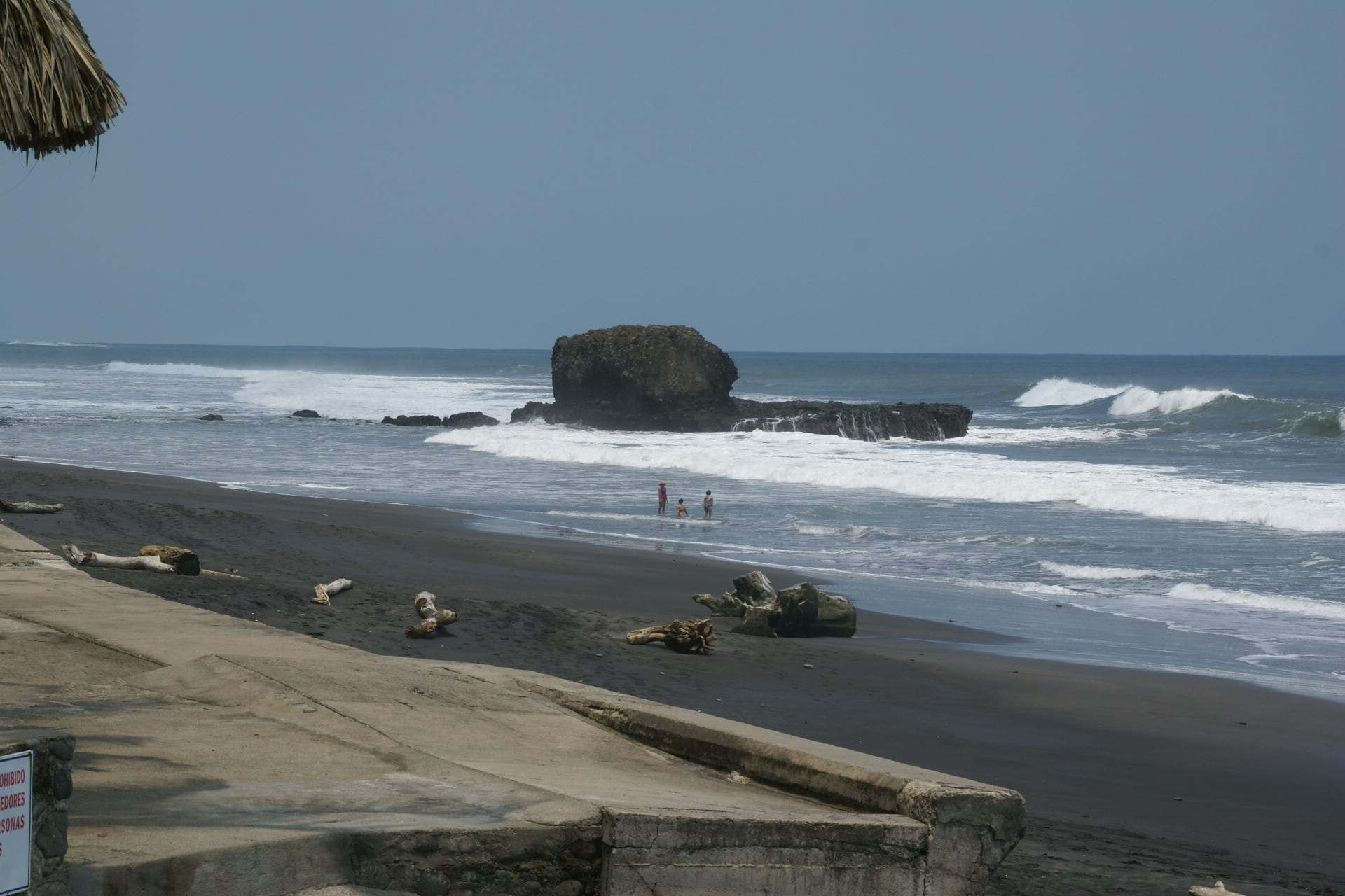 A black-sand beach with a rock offshore and waves rolling in