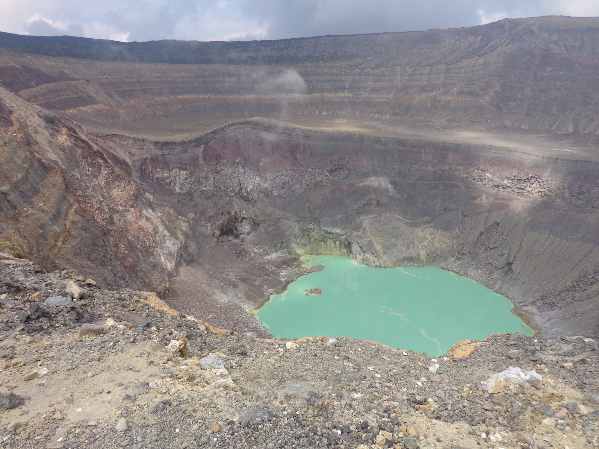 A steaming volcanic crater holding a small pale-turquoise lake, ringed by bare rock