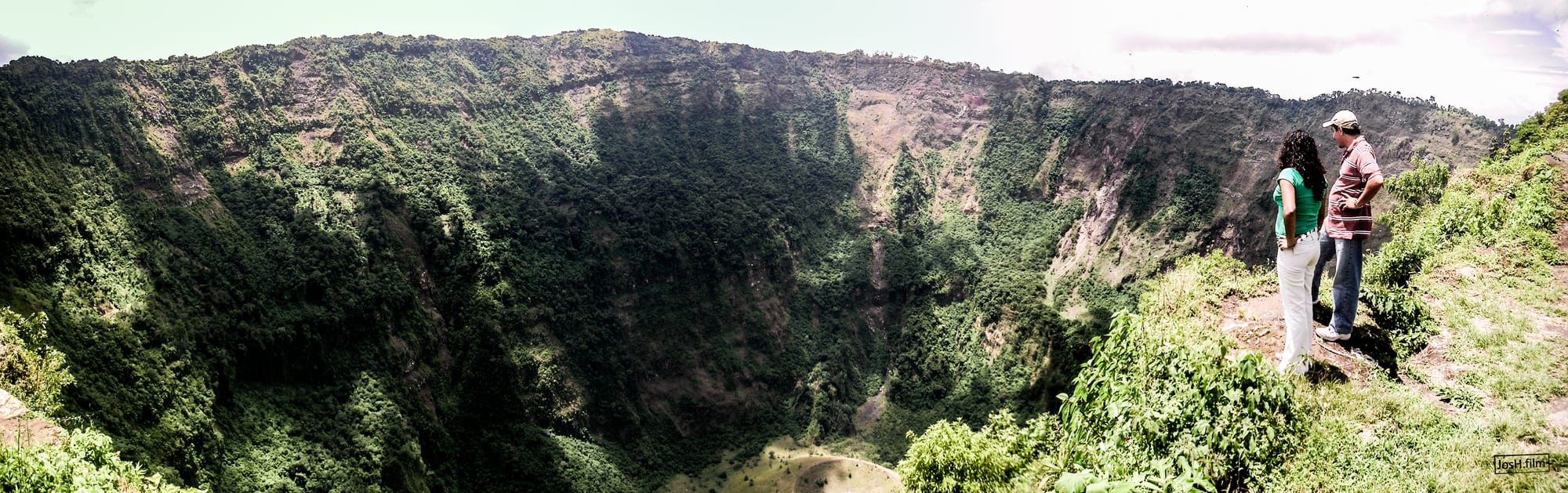 A deep, forested volcanic crater with a small cinder cone on its floor, city sprawl on the rim