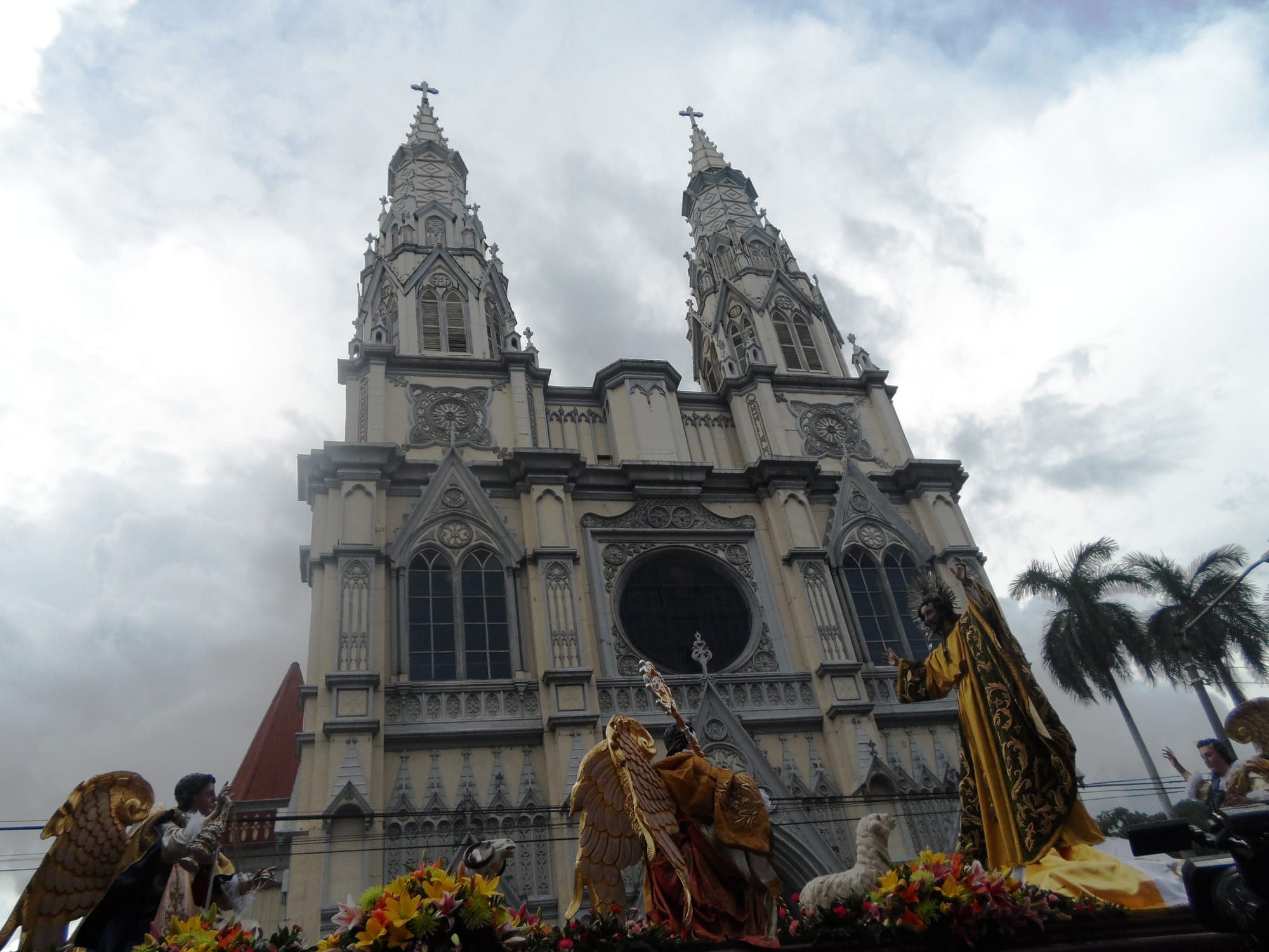 A tall white church with two ornate spires and a round window, seen from below, with gilded statues of angels and a robed figure on a flower-covered float in the foreground