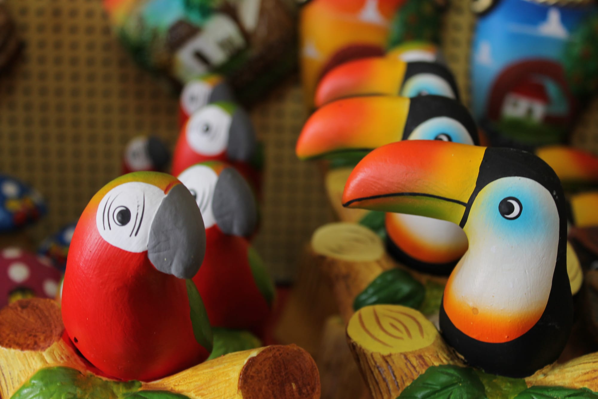 Rows of small, glossy clay figures of toucans with bright orange beaks and red parrots with grey beaks, perched on painted clay branches