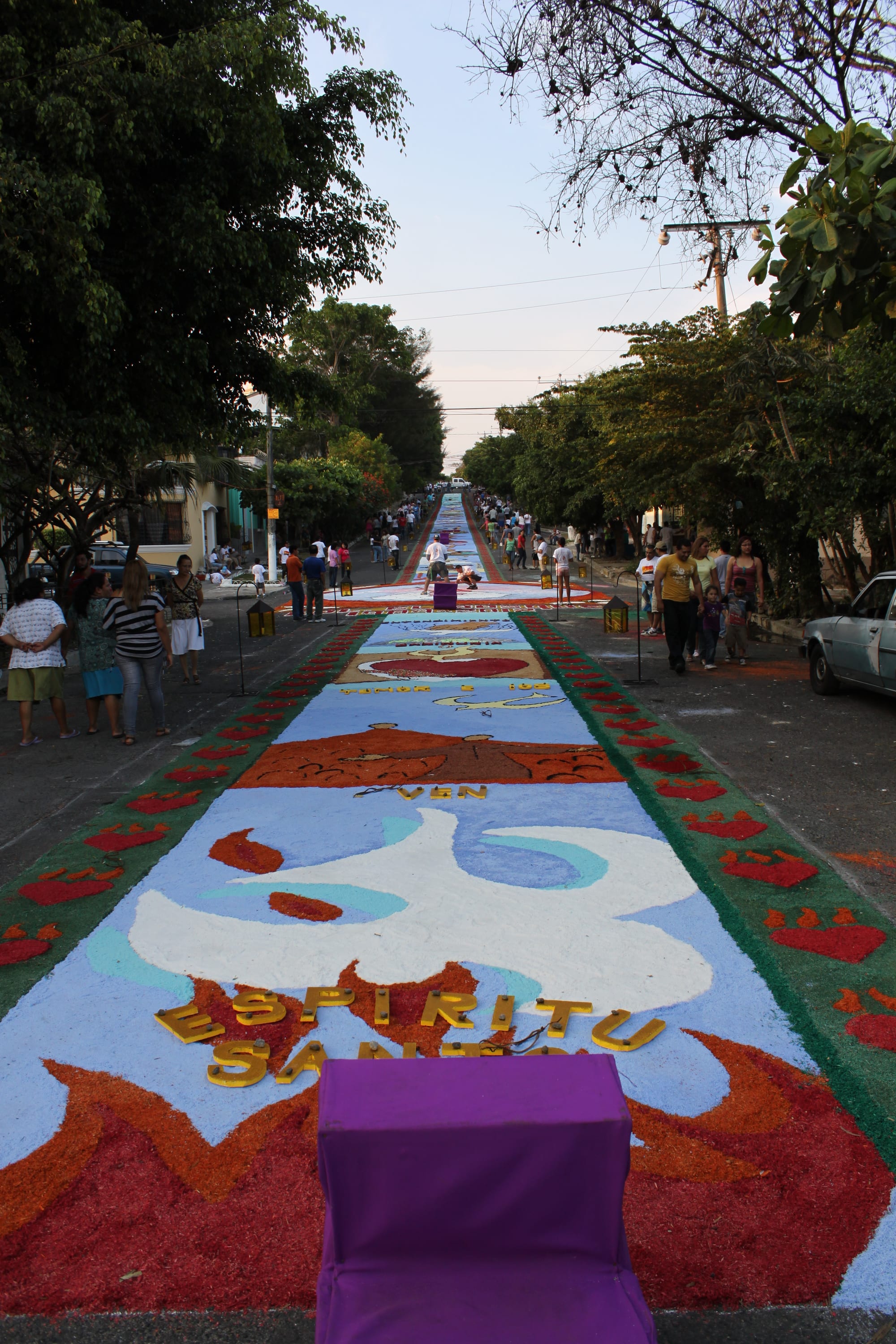 A long carpet of brightly coloured sawdust stretches down a tree-lined street, decorated with a dove, flames and the words Espíritu Santo, with people walking alongside
