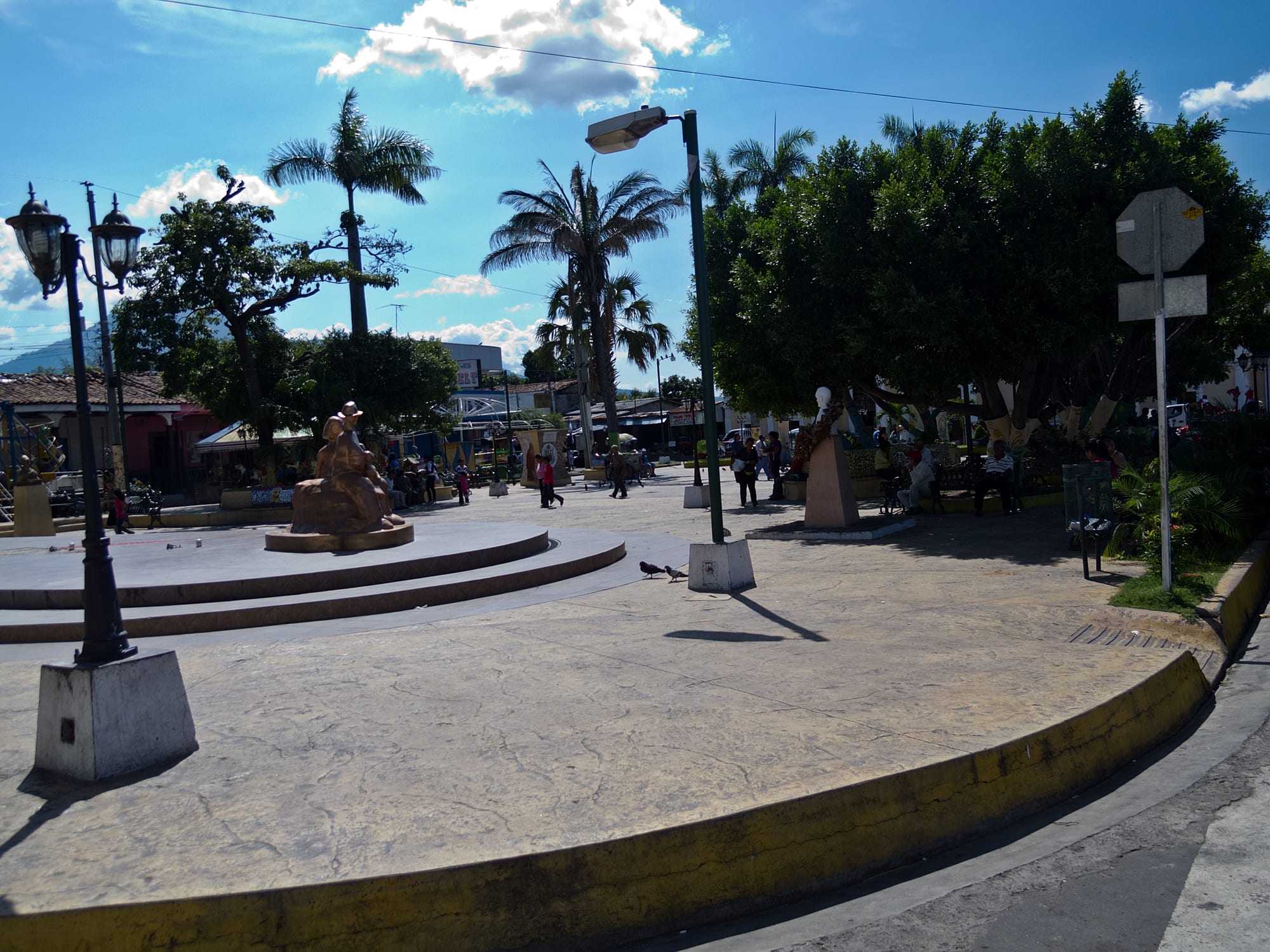 A sunny town square with palm trees, a statue and benches, with people sitting and walking about on a paved plaza