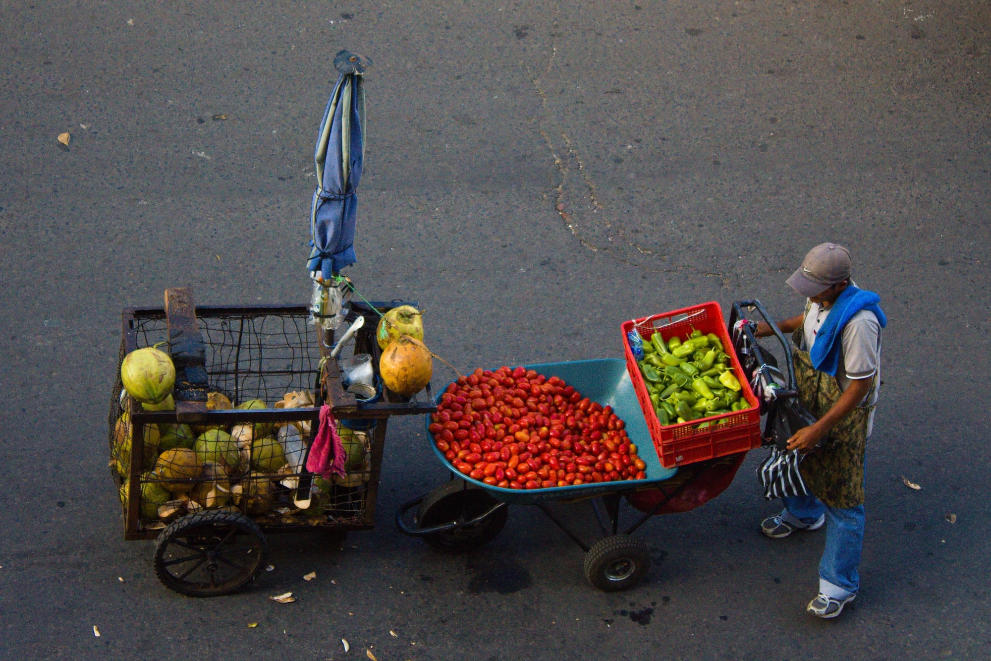 A street vendor pushing a wheeled cart loaded with coconuts, tomatoes and green peppers along a street