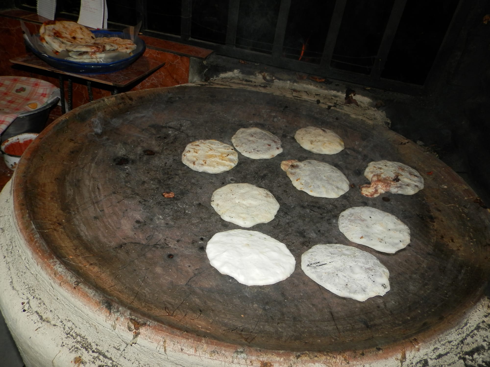 Nine pupusas cooking on a large blackened clay griddle, with a plate of finished pupusas visible in the background