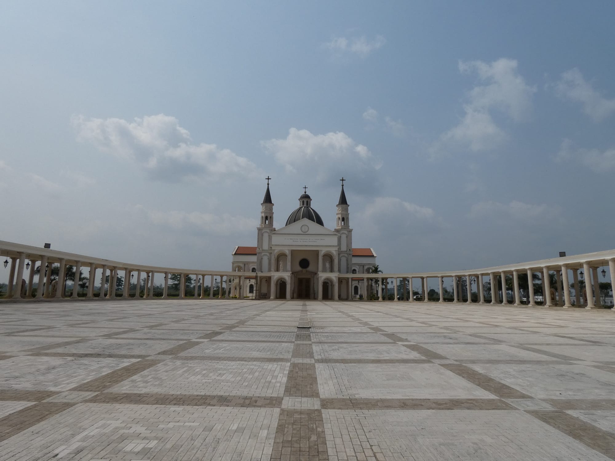A white basilica with a central dome and two slender towers topped by crosses stands at the far end of a vast paved plaza enclosed by curving white colonnades, under a cloudy sky