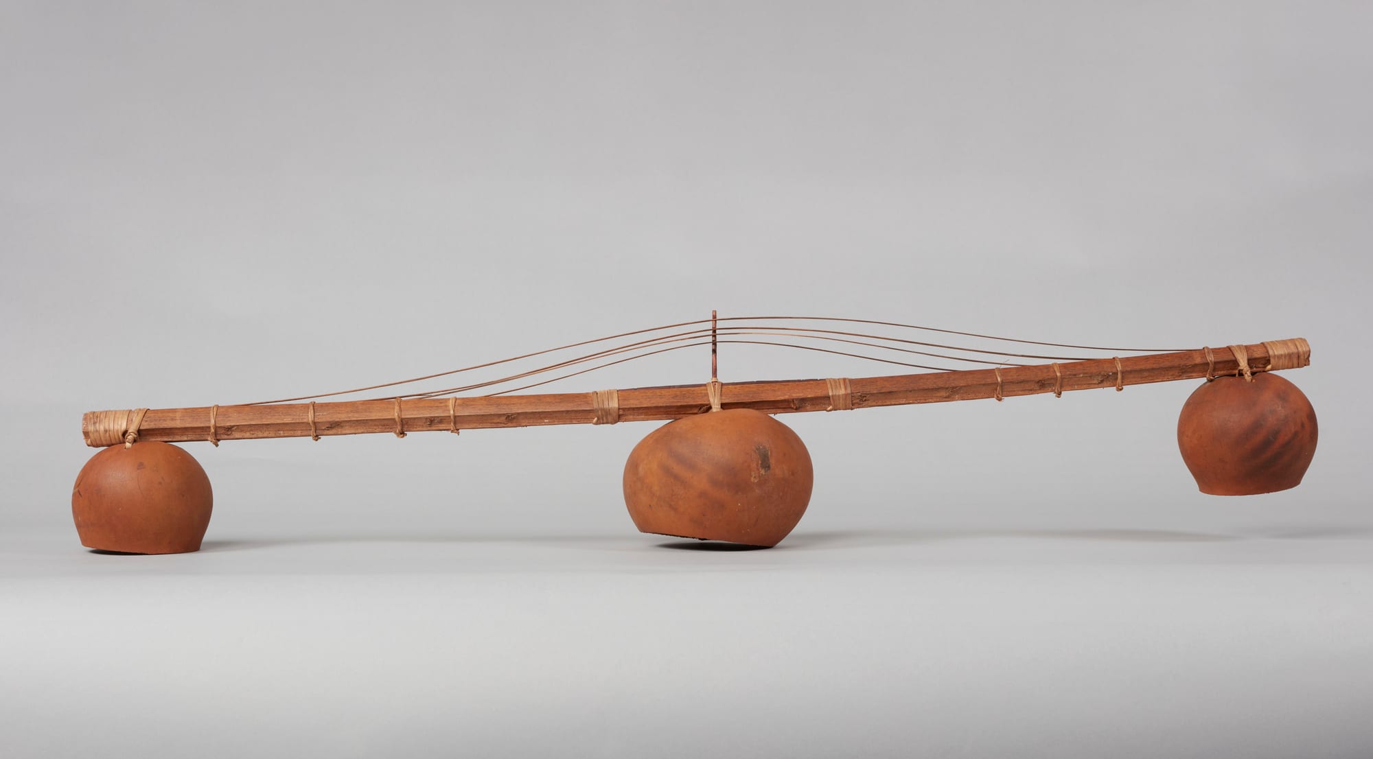 A long, straight wooden stem bound with fibre, with thin strings raised over a small upright bridge at its centre and three round brown gourds hanging beneath it, photographed against a plain grey background