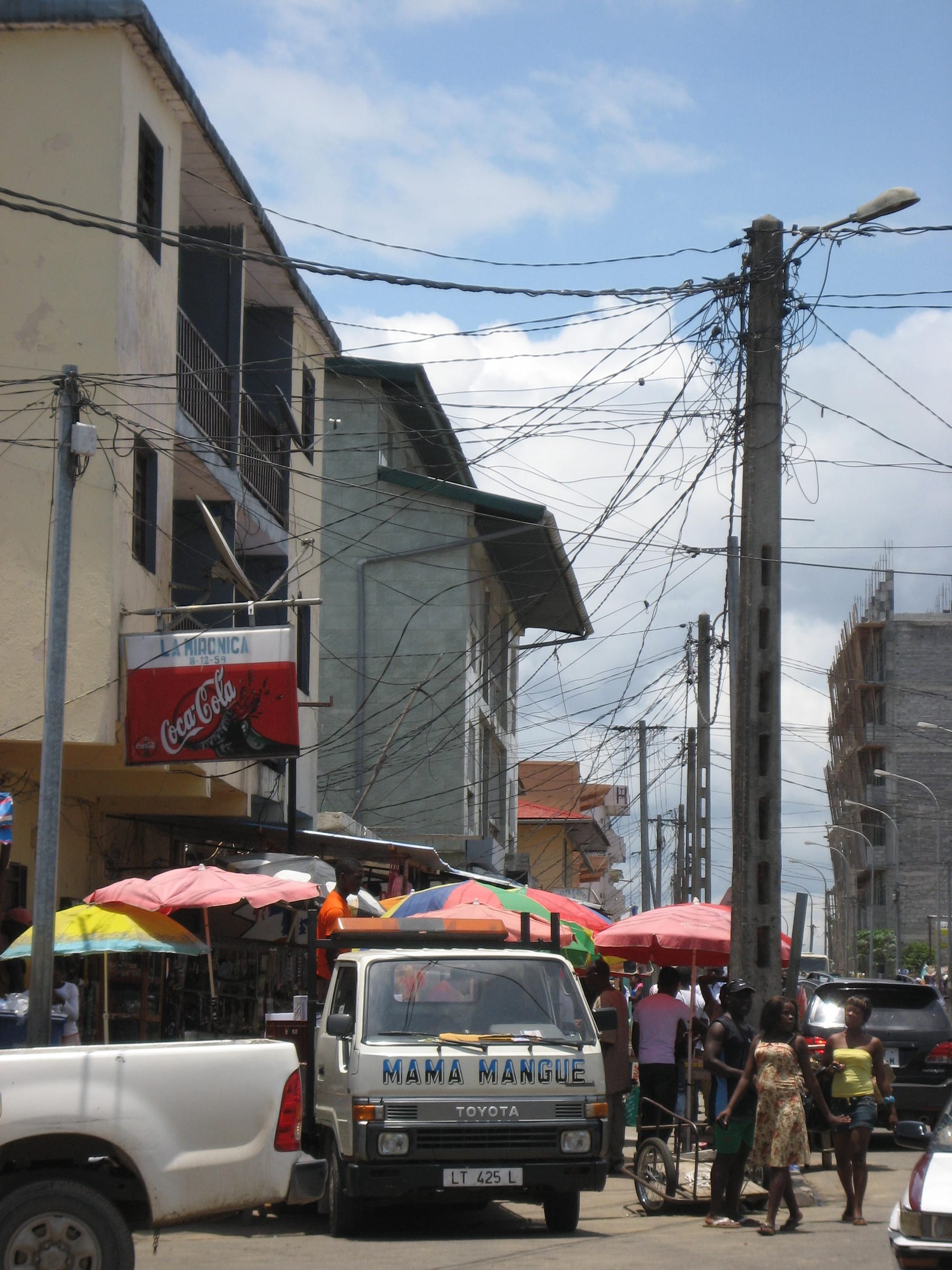 A busy street market in Bata with colourful umbrellas over stalls, a parked truck, and people walking between tangled overhead wires