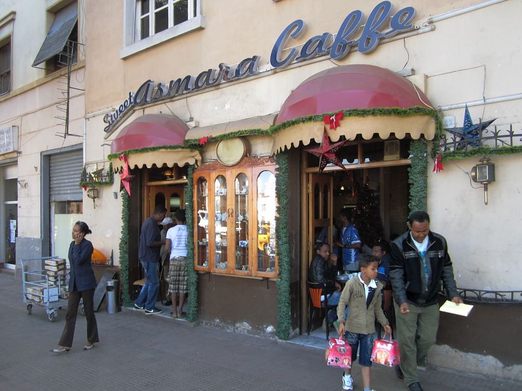The pink-awninged storefront of Sweet Asmara Caffe on a street in Asmara, with people walking past and customers visible inside