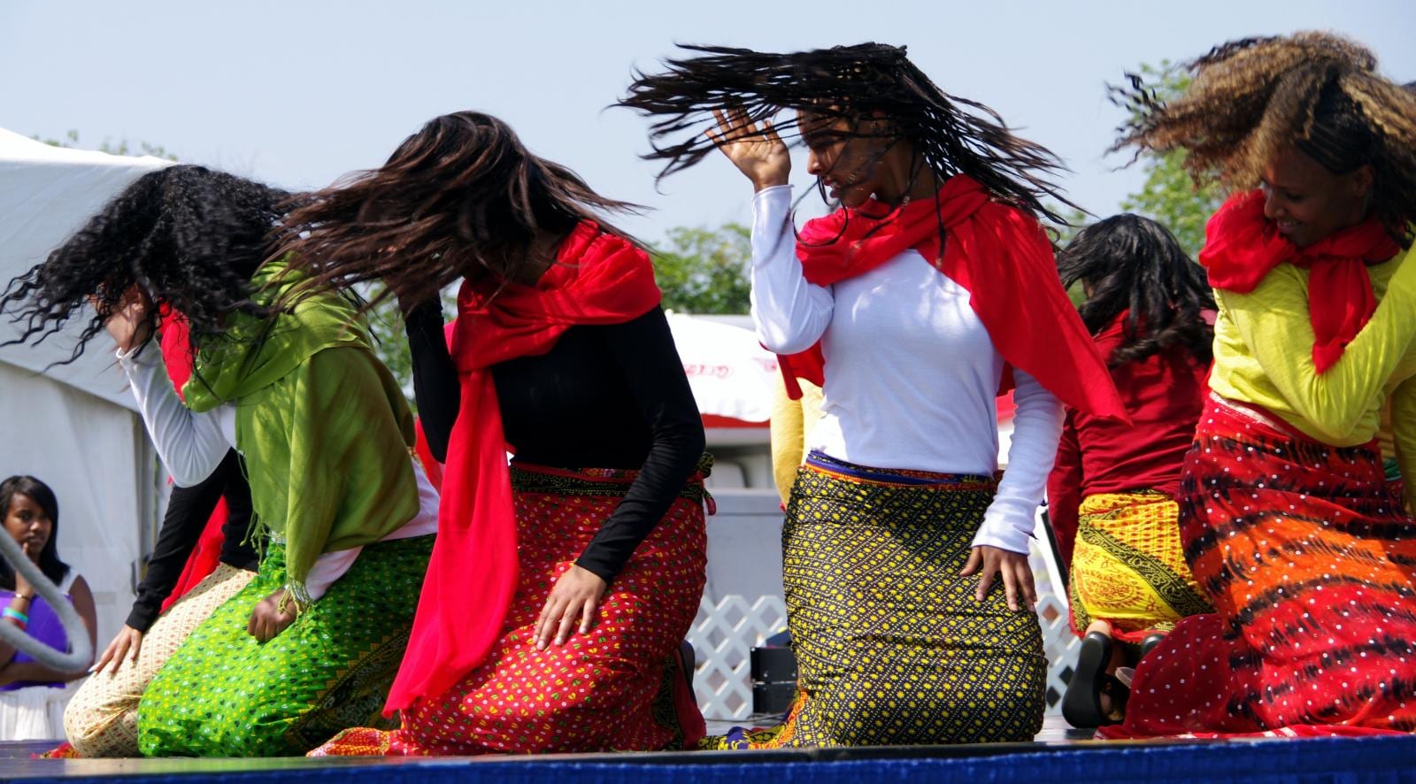 A row of women in red scarves, colourful long skirts and long-sleeved tops kneel on a stage and toss their long hair as they dance, with a white tent behind