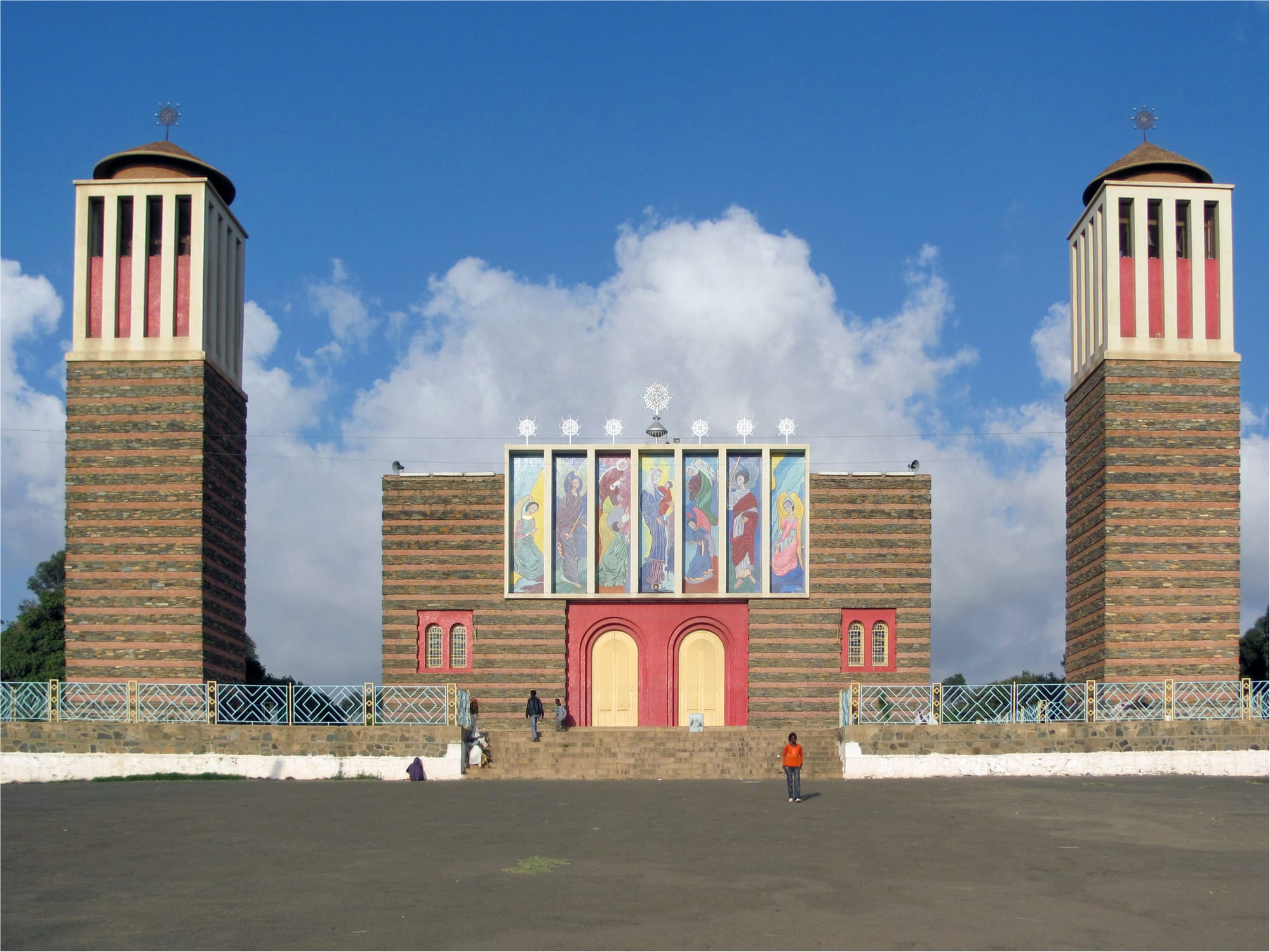 A church with two tall towers of banded stone either side of a central facade painted with a row of saints above red arched doors, set back behind a broad open square under a blue sky with clouds