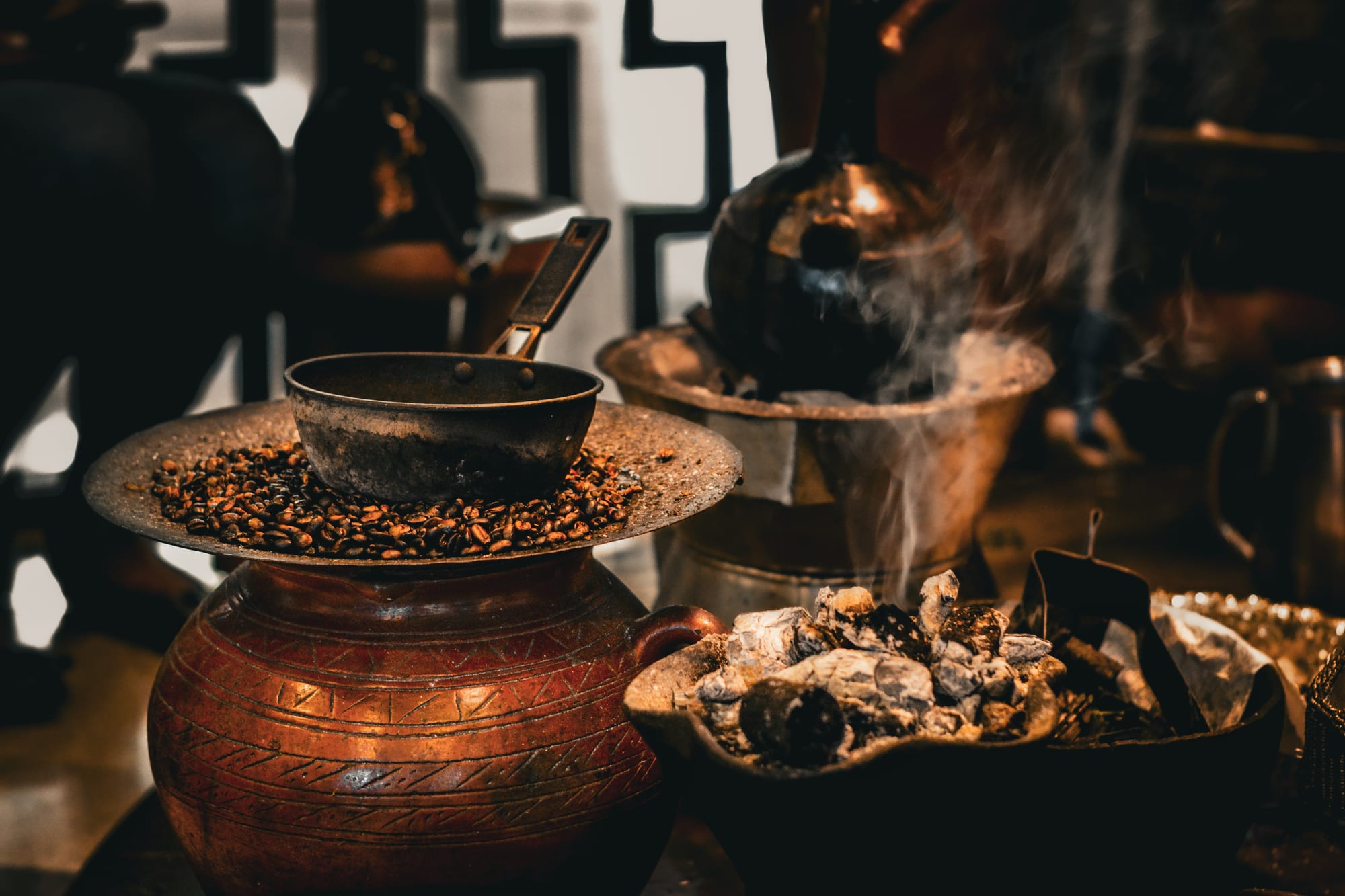 A dark metal pan on a flat tray of coffee beans rests on a decorated clay pot, beside a smoking brazier of charcoal and a black clay coffee pot in the background