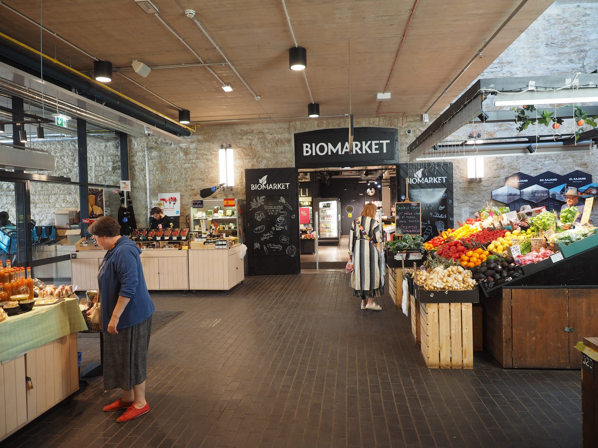 Fresh fruit and vegetable stalls at the covered Balti Jaama Turg market hall in Tallinn, with a shopper browsing the produce