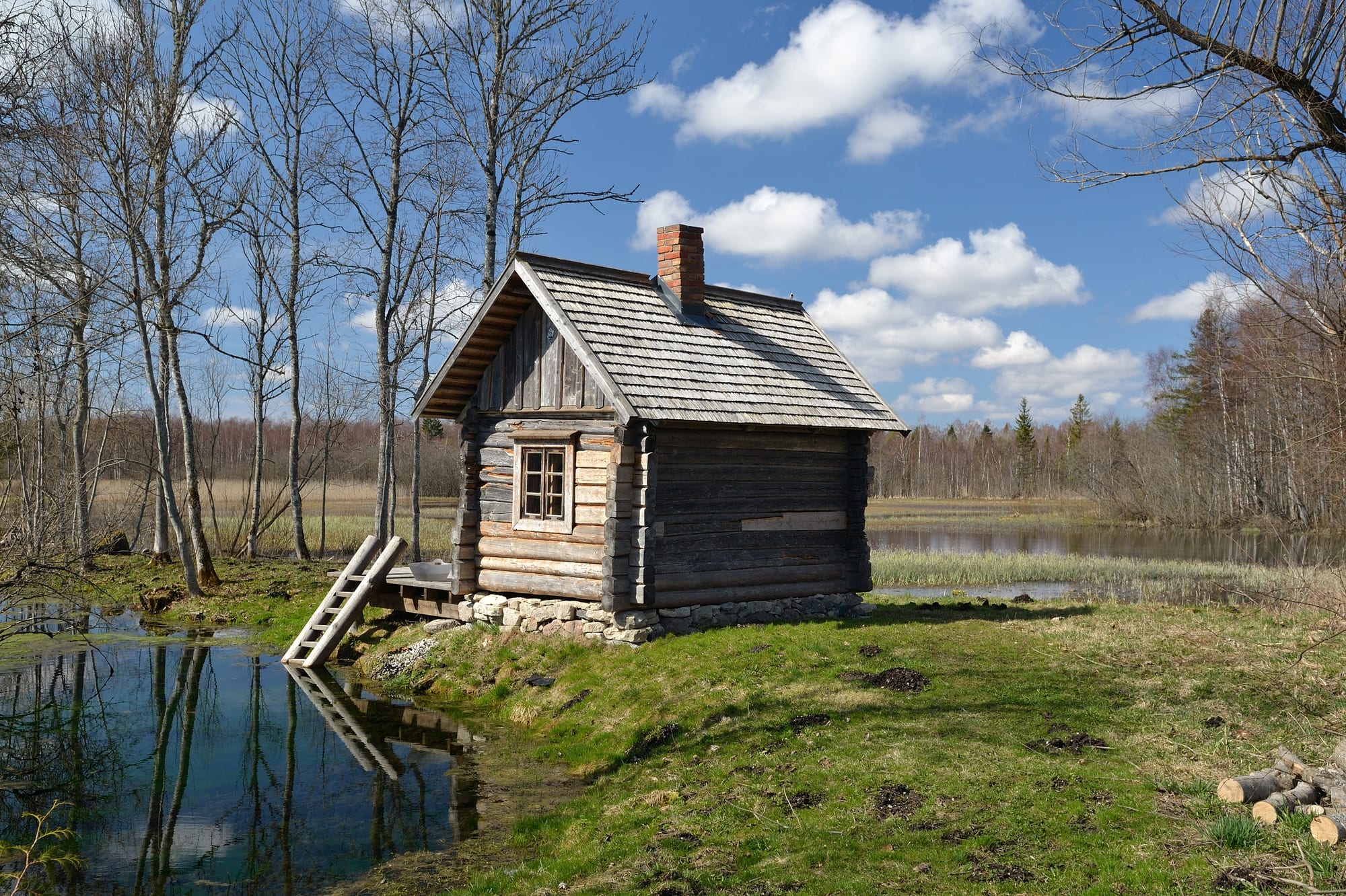 A small wooden sauna cabin with a stone chimney standing beside a still spring lake, reflected in the water