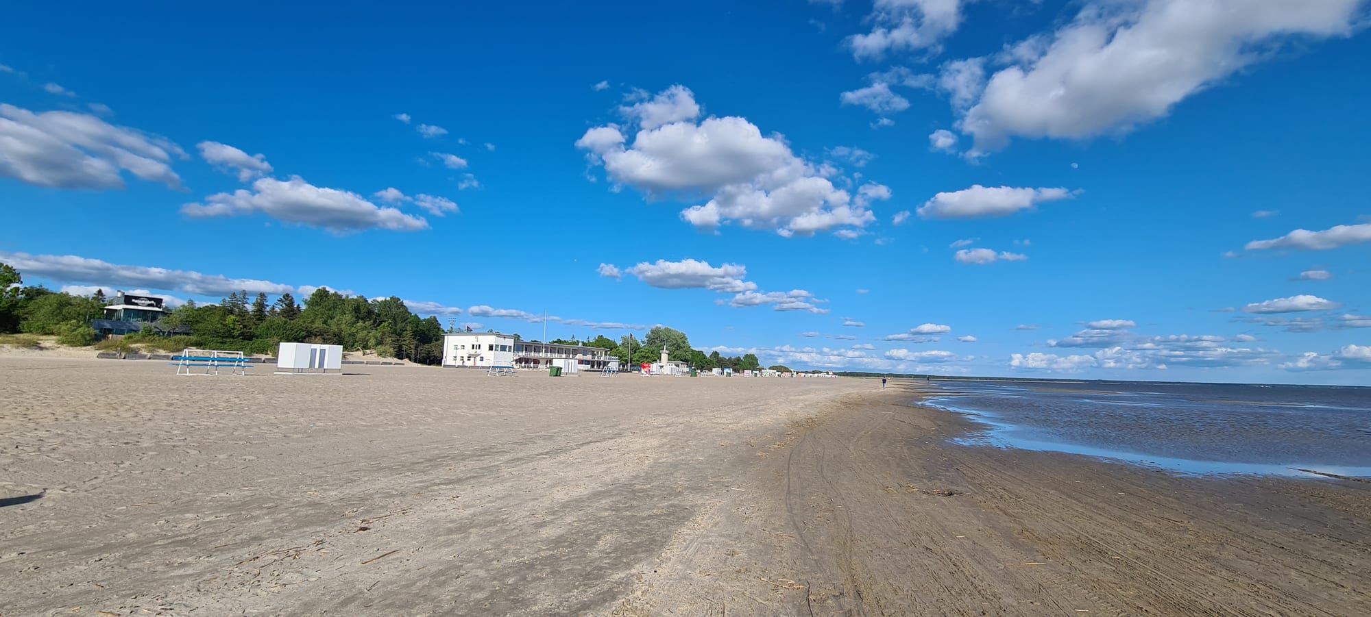 A wide, shallow-sloping sandy beach with a few people, low dunes and pines behind