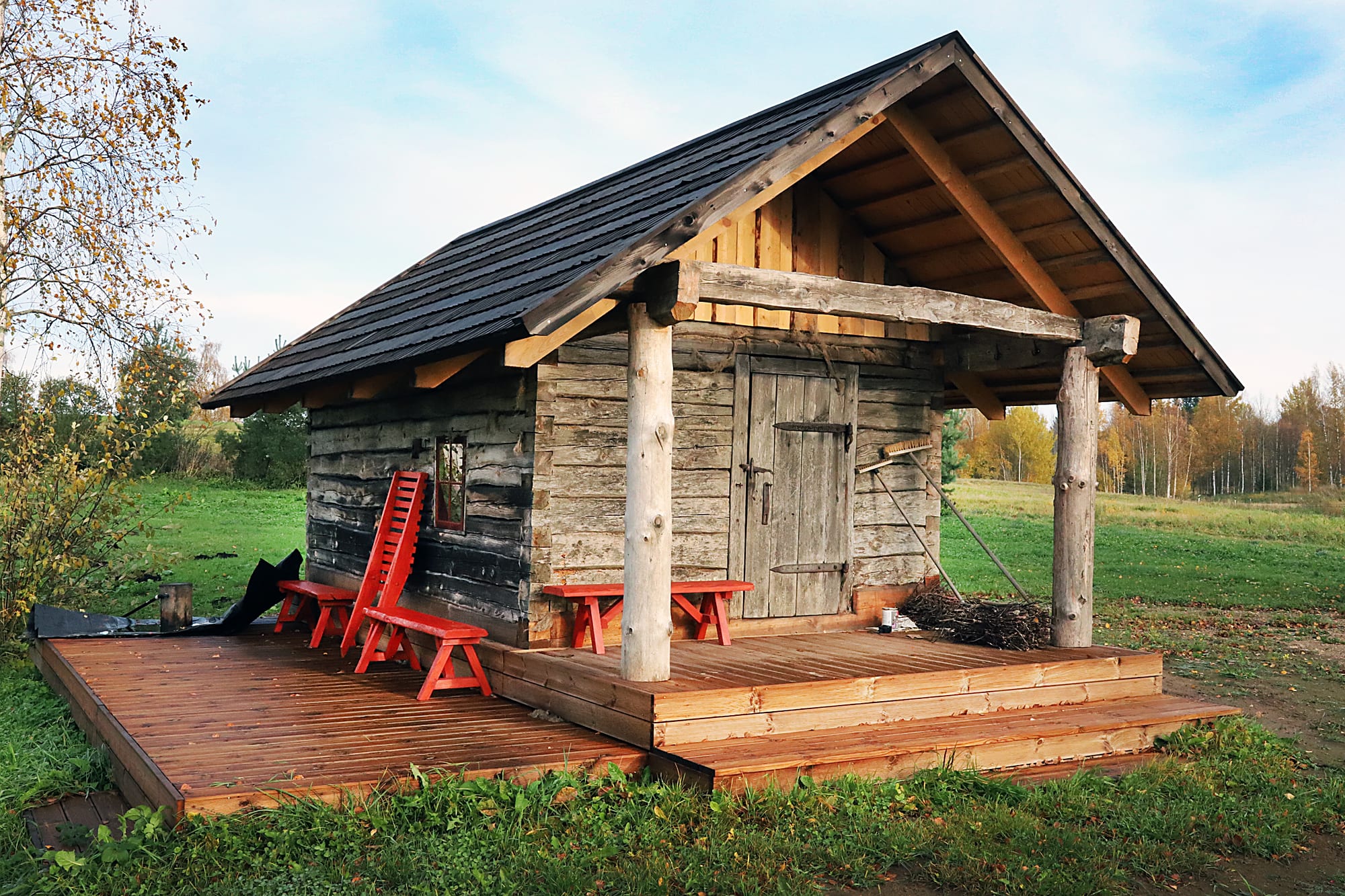 A small weathered log sauna building with a dark roof and a covered wooden porch, red wooden benches and a bundle of birch branches, standing in a green field with autumn trees behind