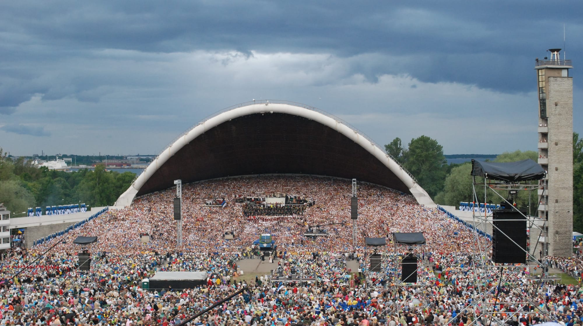 A huge white-rimmed arched stage filled with thousands of singers under a stormy grey sky, with a vast crowd spread across the ground in front and a tall stone tower to the right