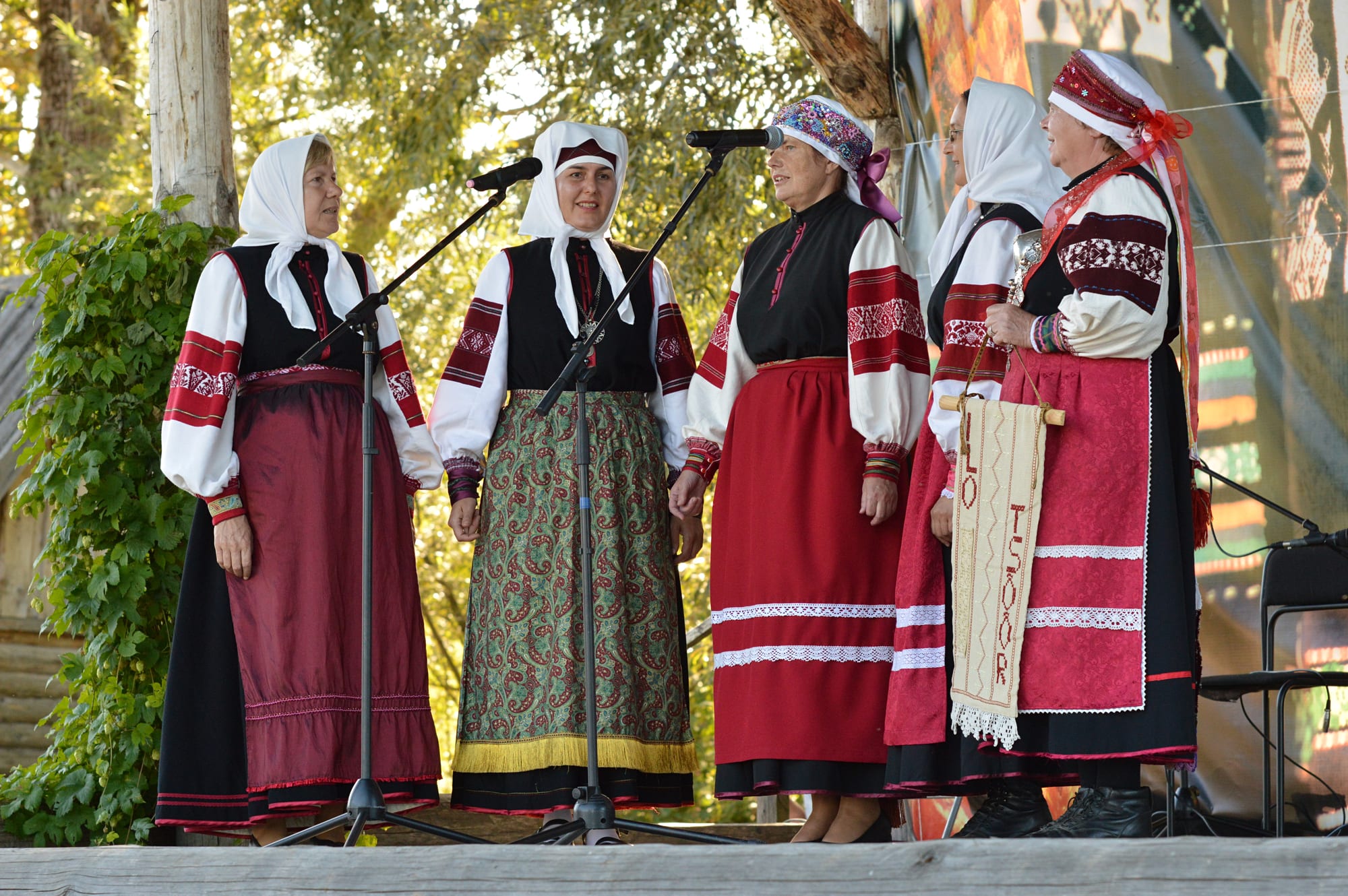 Five women in traditional Seto dress with white headscarves, embroidered white blouses and long red and dark skirts stand singing at microphones on an outdoor wooden stage