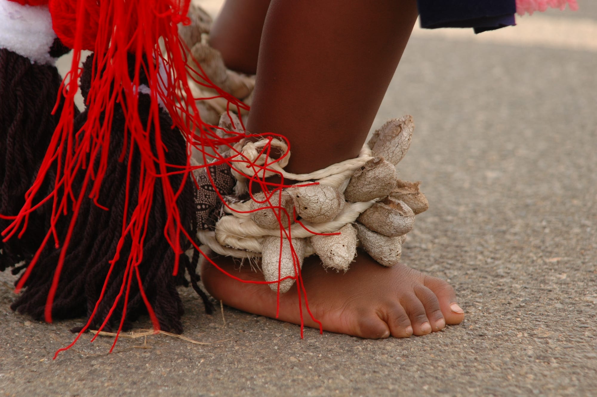 Close-up of a dancer's bare foot on tarmac, the ankle wrapped in a rattle of dried pods, beside red and dark wool tassels