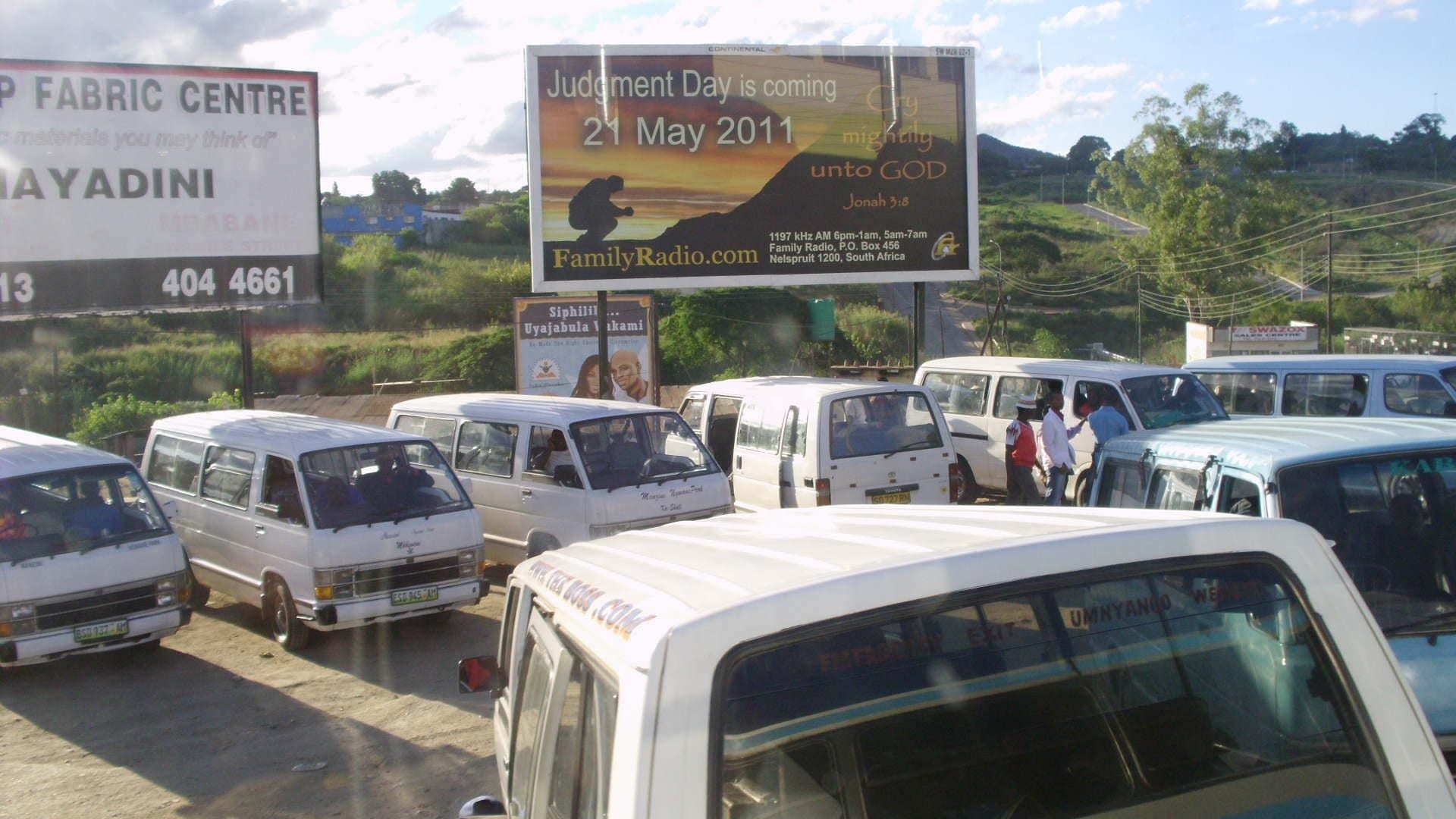A row of white minibus taxis parked at a taxi rank in Eswatini, with billboards and hills behind