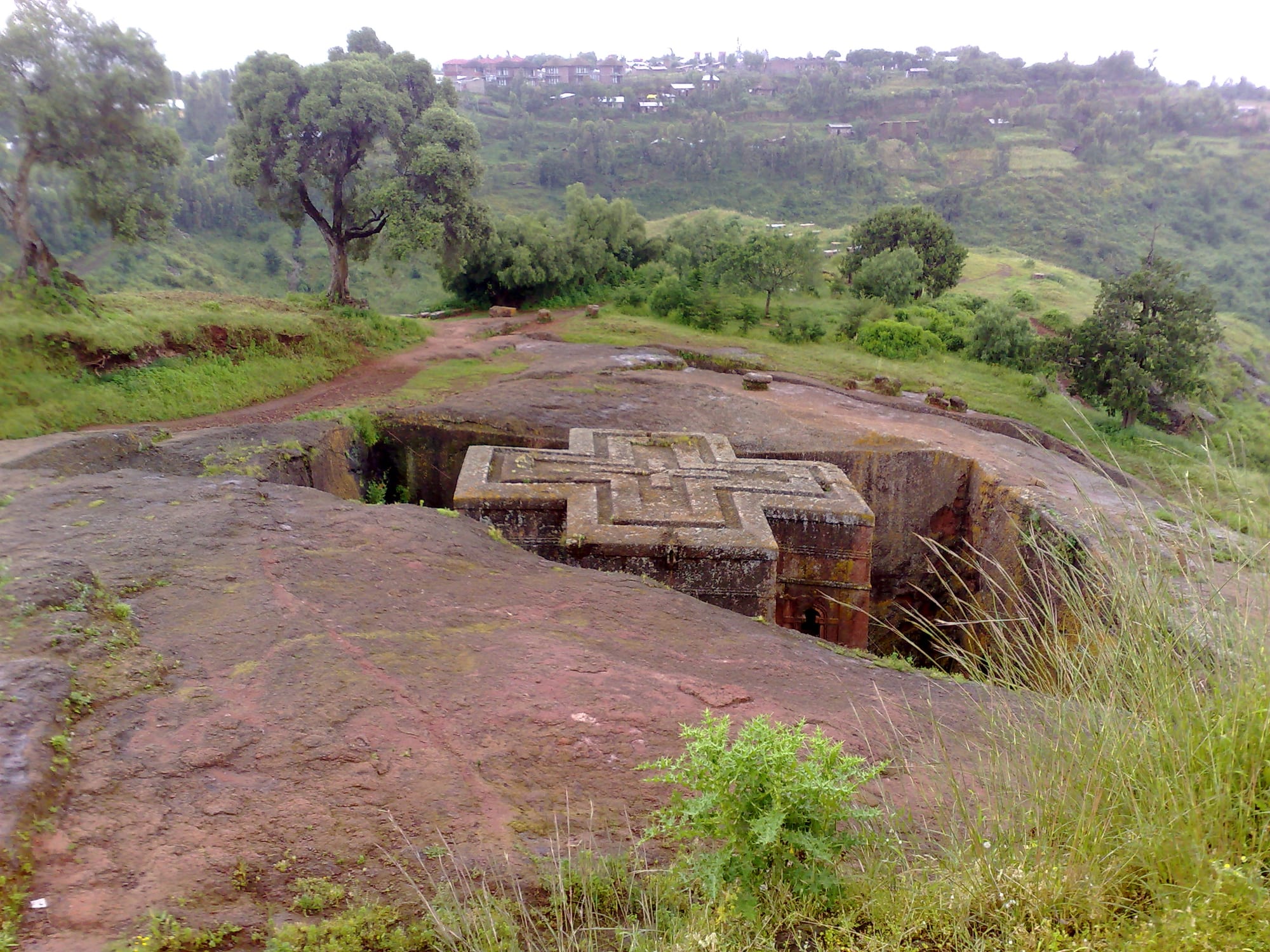 A cross-shaped church carved down into pink rock, isolated at the bottom of a deep pit