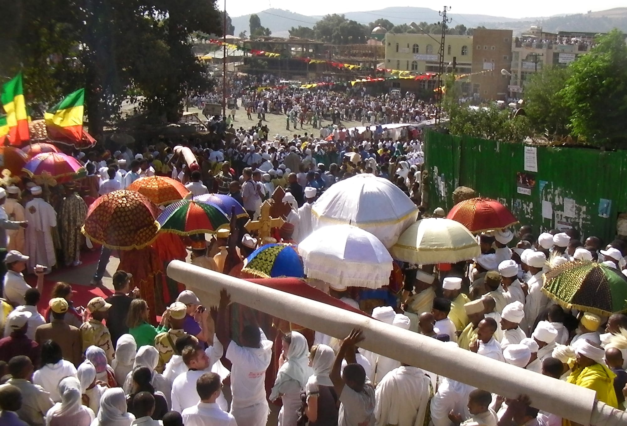 A large crowd dressed mostly in white surrounds priests walking under brightly coloured fringed umbrellas, with processional crosses held aloft and Ethiopian flags at the left