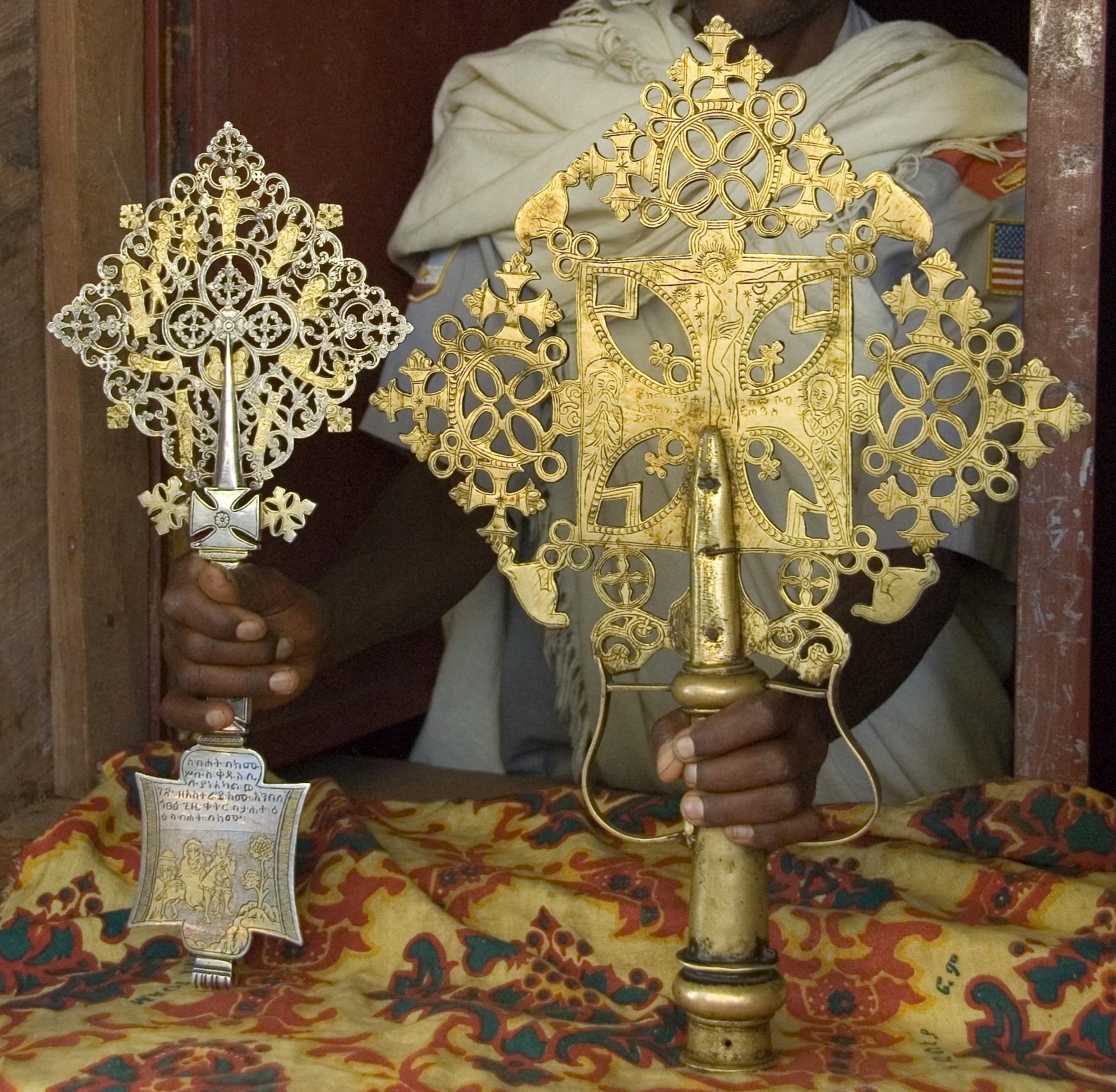 Two ornate metal crosses with lace-like openwork, one silver and one gold-coloured, held upright over a patterned cloth by a man in a white shawl