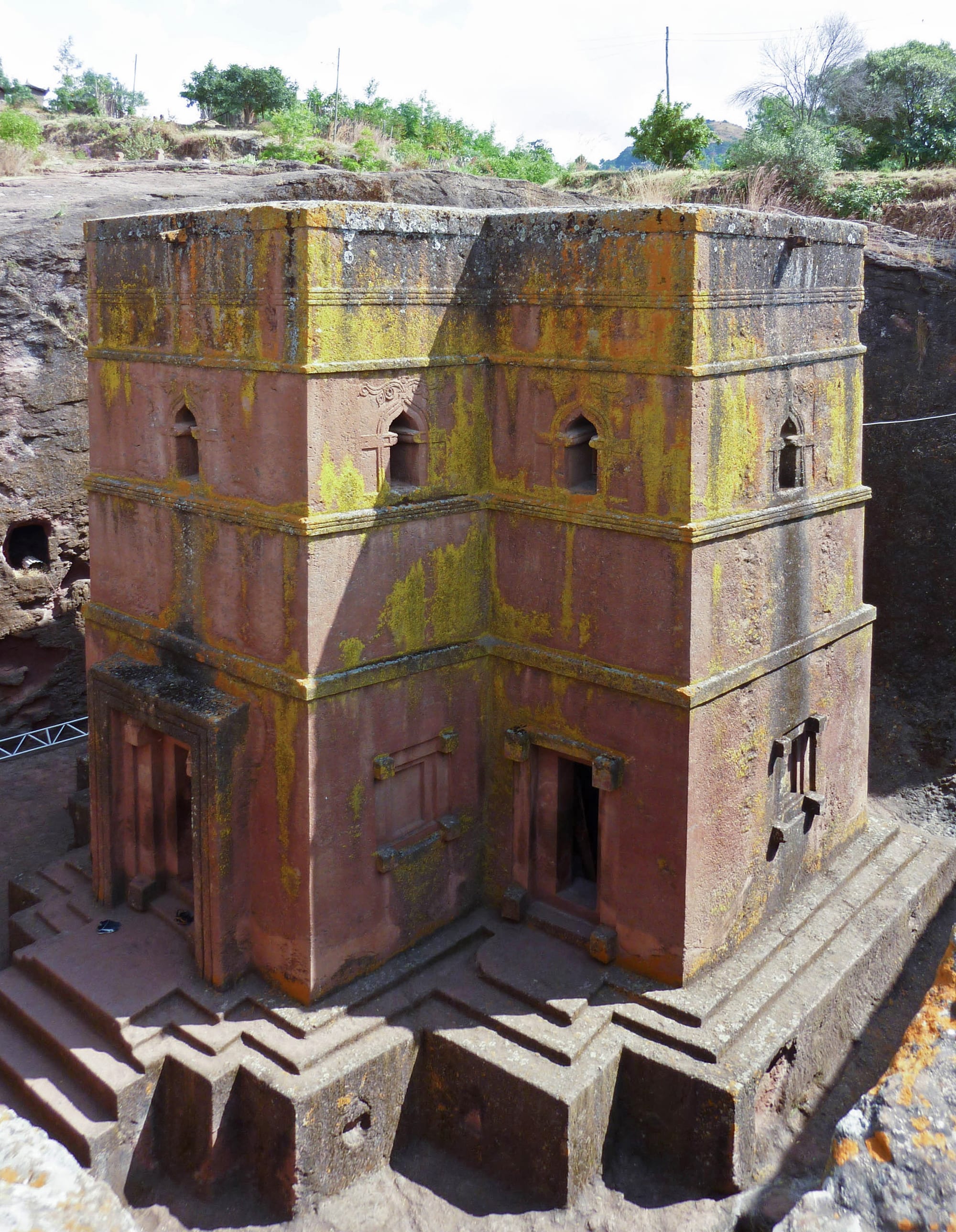 A cross-shaped church carved down into solid red rock, seen from ground level above