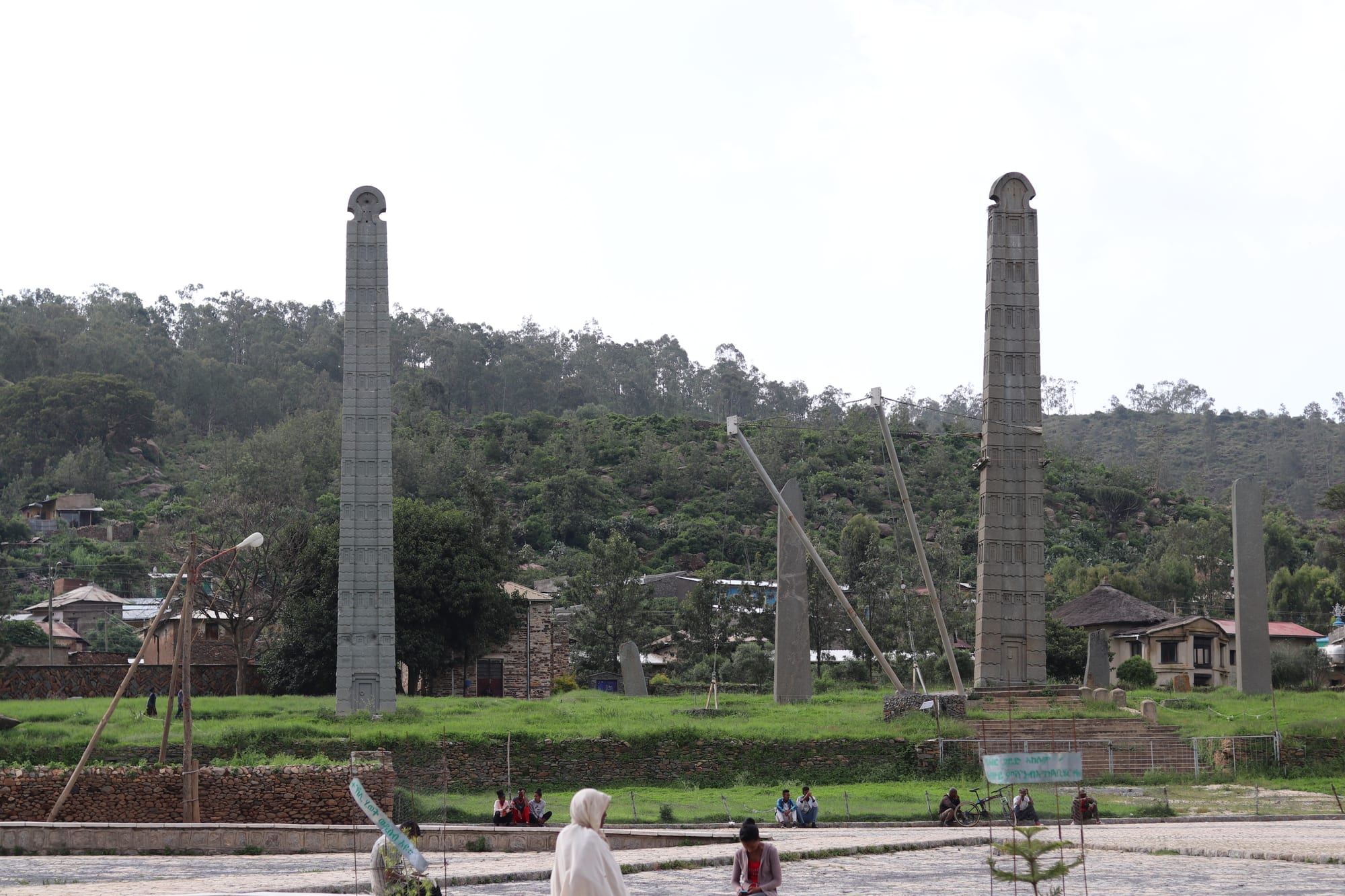 A tall, narrow carved granite stele standing among ruins under a blue sky