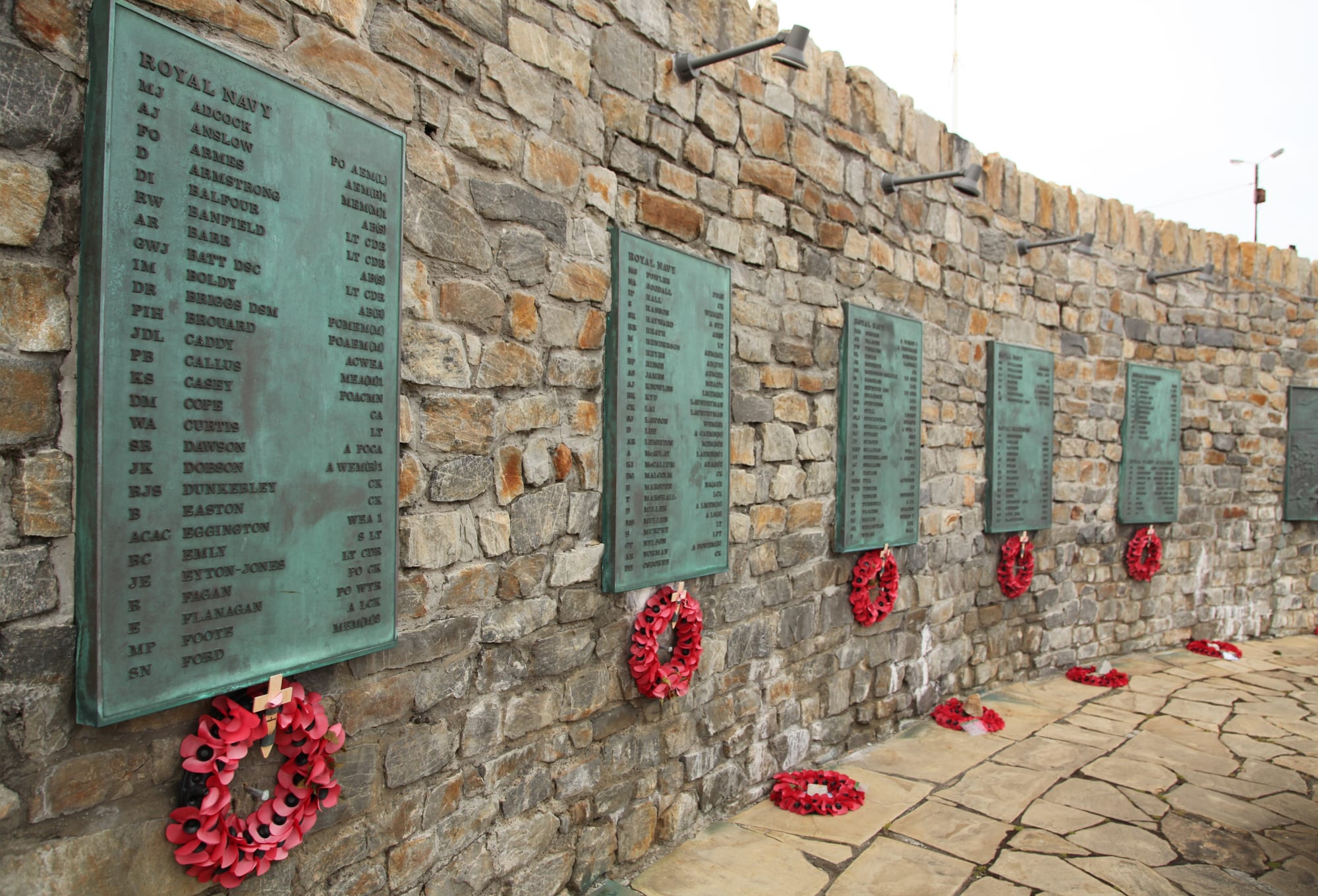 A long stone wall lined with green bronze plaques listing names, with red poppy wreaths hanging below each plaque and more wreaths laid on the paving