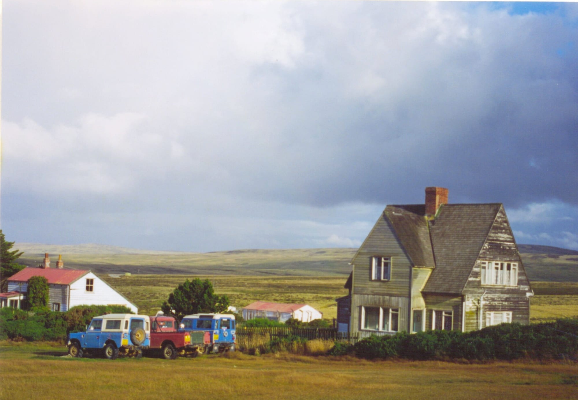 A weathered two-storey wooden farmhouse with a brick chimney beside three old Land Rovers parked on the grass, with a white cottage and open moorland stretching to low hills under a heavy sky