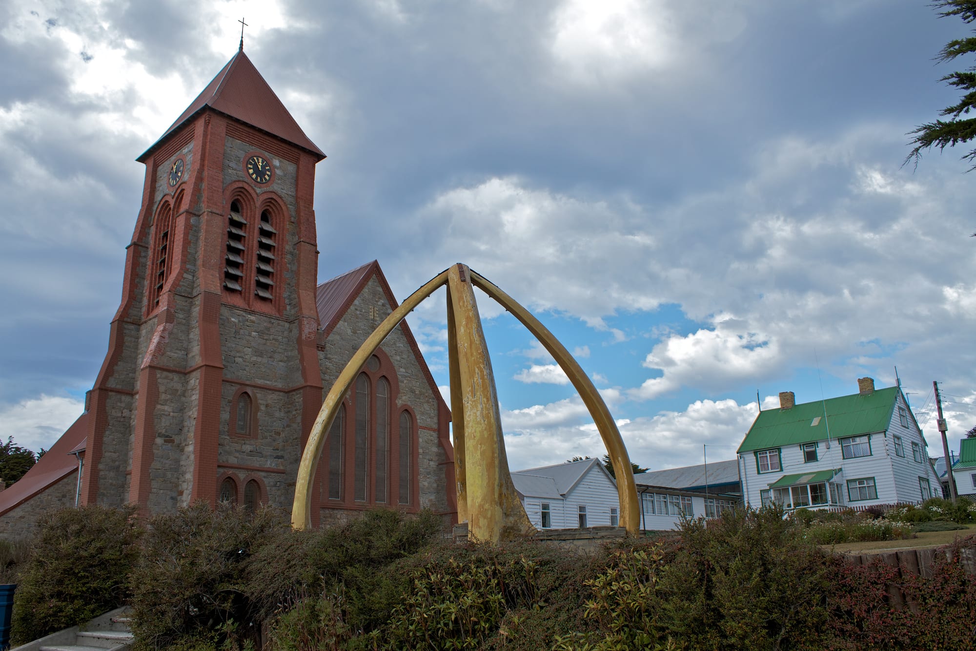 A stone and red-brick church with a tall square clock tower beside an arch made of two huge pale whale jawbones, with white houses and a cloudy sky beyond
