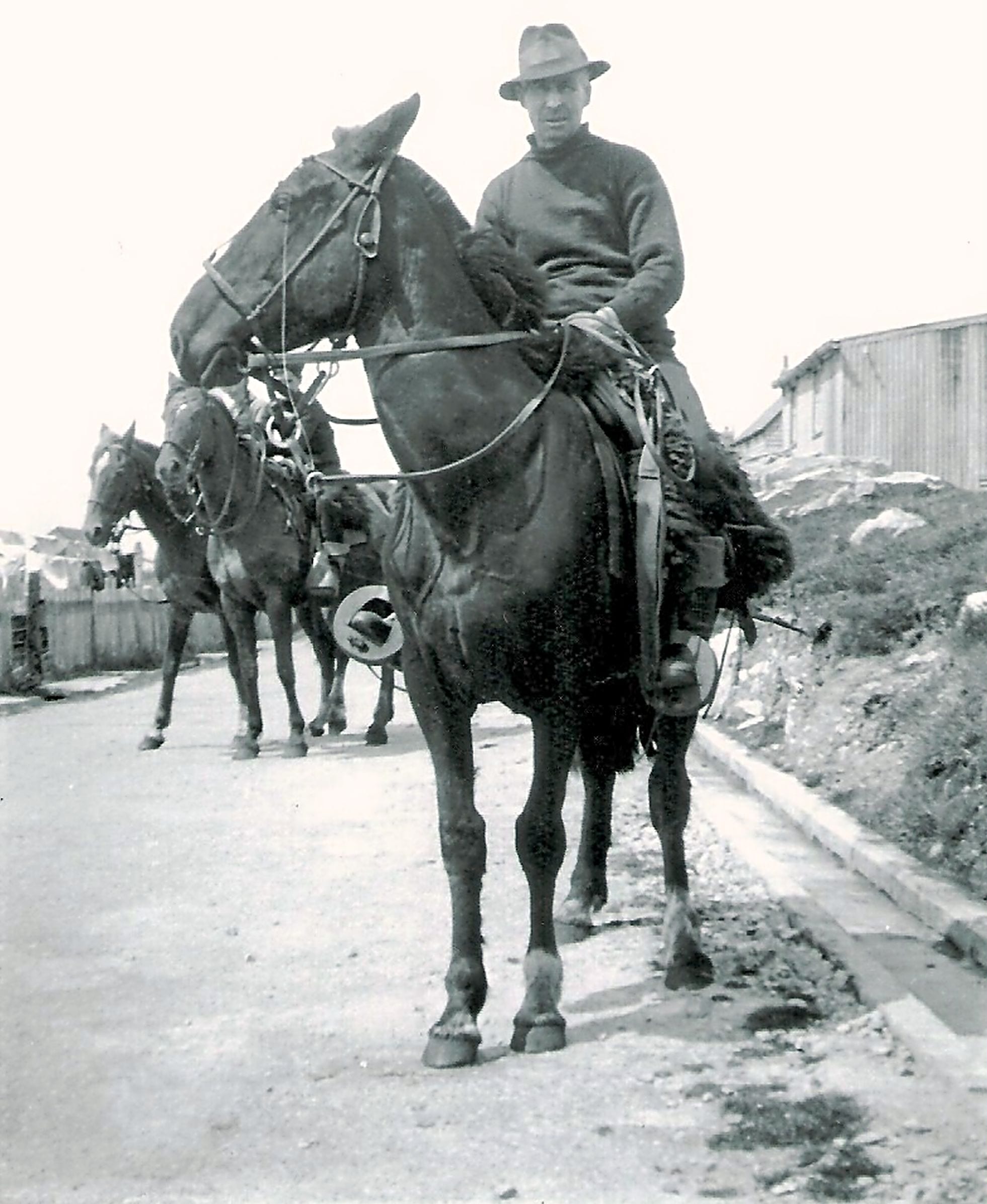 A black and white photograph of a man in a hat and heavy jumper sitting on a dark horse with a sheepskin-covered saddle and leather tack, with two more saddled horses behind him on a track beside a timber building