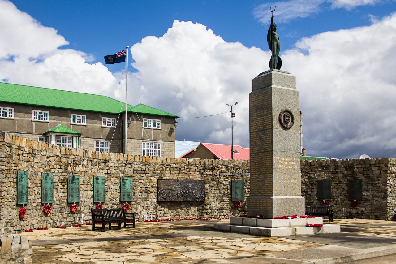 A pale stone memorial with a domed top and bronze plaques, on a lawn by the sea in Stanley