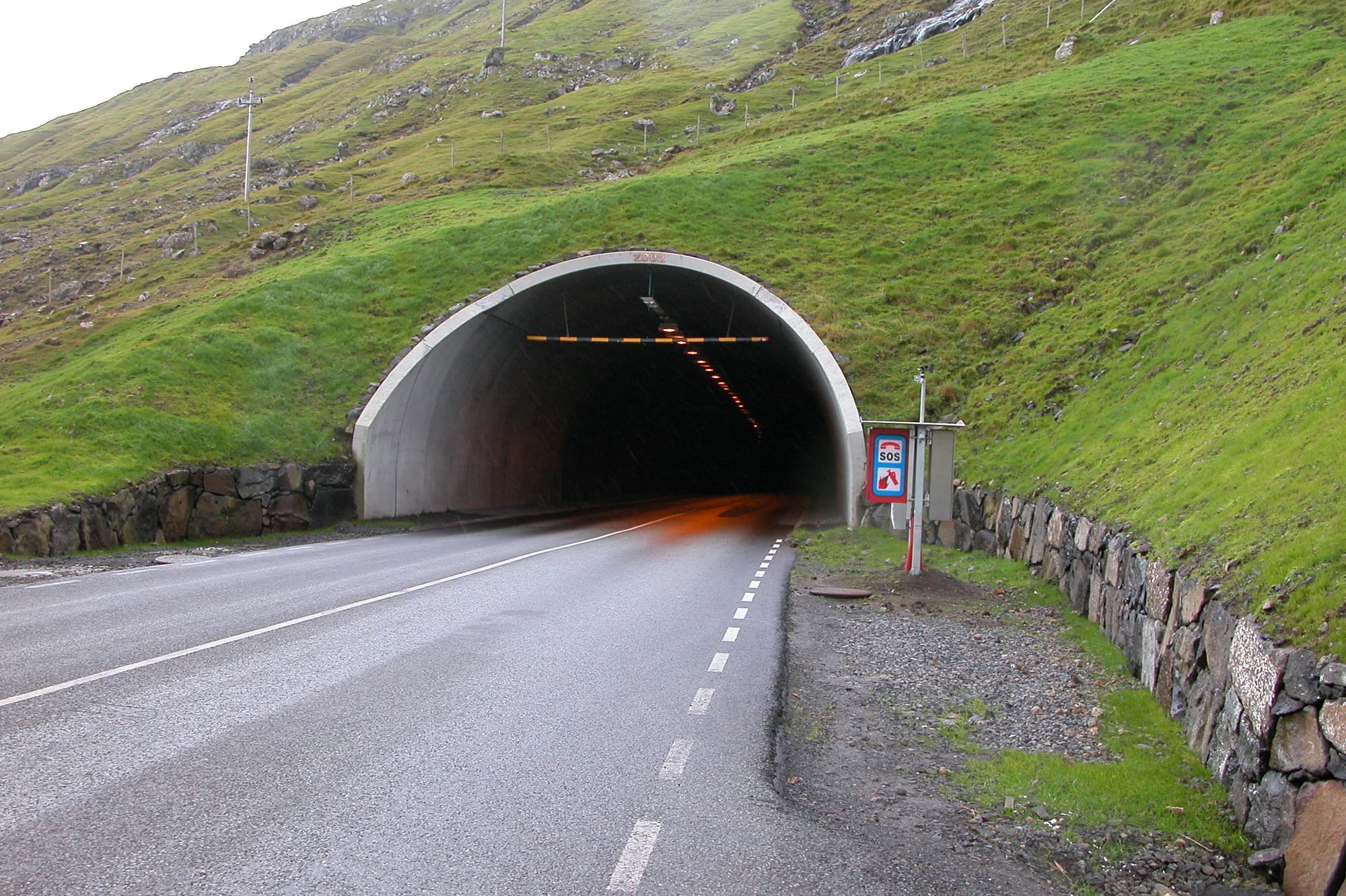 The entrance to a road tunnel cut into a steep green hillside in the Faroe Islands, with a wet road leading into it
