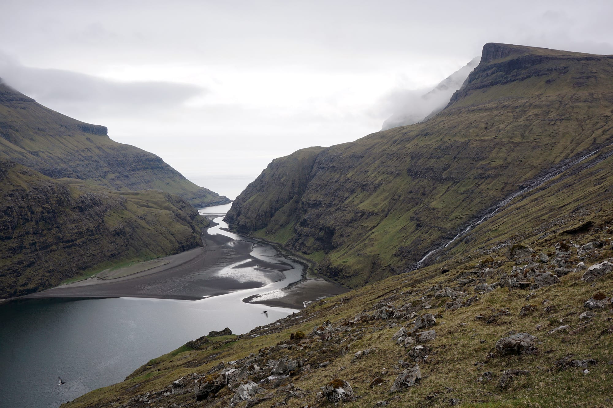 A lagoon and turf-roofed farm buildings surrounded by very steep green mountainsides