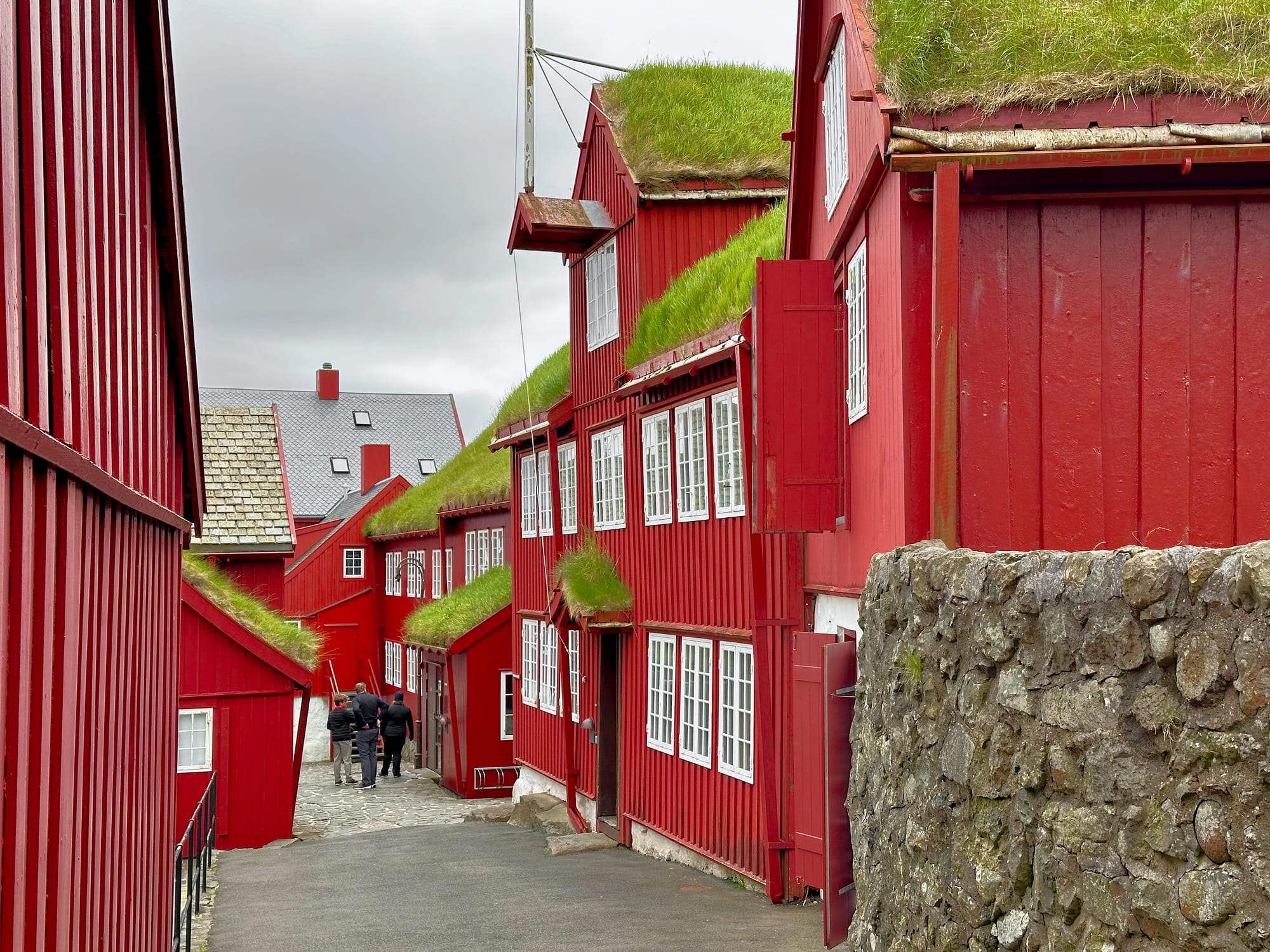 A cluster of small red-painted timber buildings with turf roofs on a rocky harbour promontory