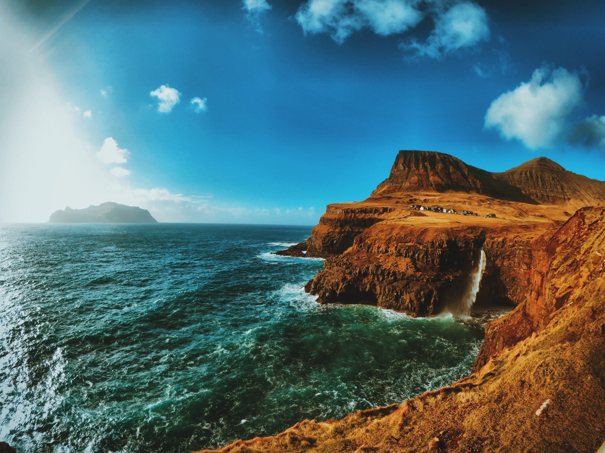 A single narrow waterfall dropping off a green cliff edge into the sea, a village behind