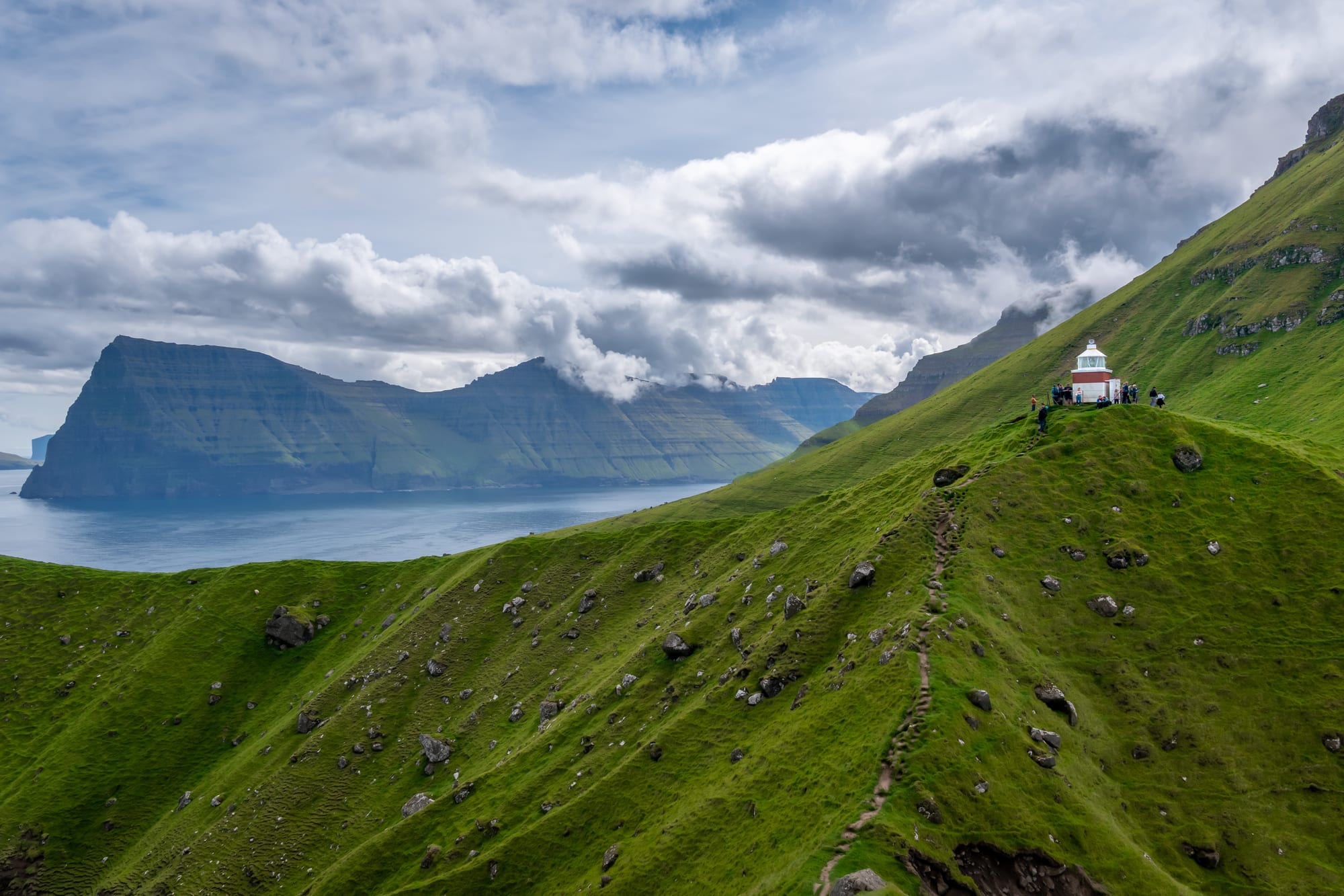 A tiny white lighthouse on a knife-edge green ridge, sheer cliffs falling to the sea on both sides