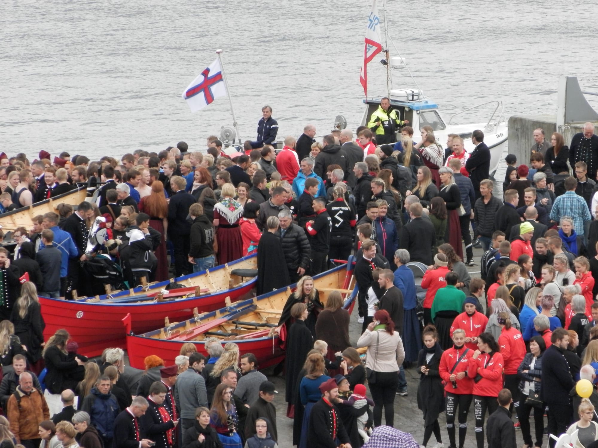 A large crowd, some in Faroese national dress, gathered on a harbour front around red wooden rowing boats, with the red, blue and white Faroese flag flying above and grey water beyond